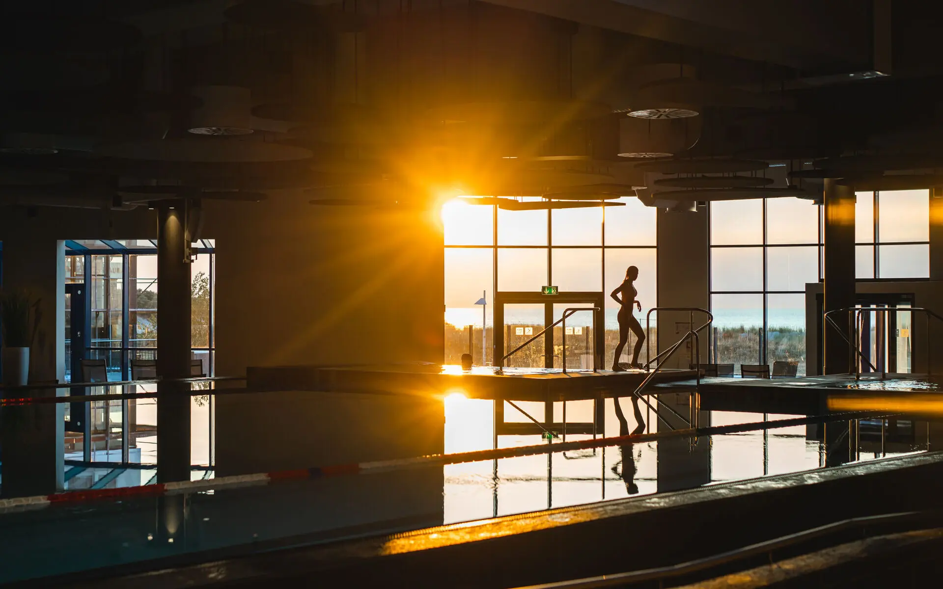 Innenpool aja Warnemünde Gegen die Abendsonne fotografiert sieht man den Innenpoolberich im aja Warnemünde vor einer großen Fensterfront mit Aussicht auf die Strandlandschaft. Im Schatten erkennt man eine Frau, die hinten am Beckenrand entlang geht.