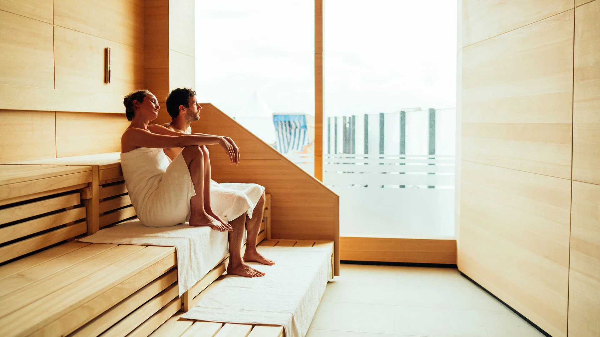 Zwei Menschen sitzen in Handtücher gewickelt in einer Sauna und entspannen.