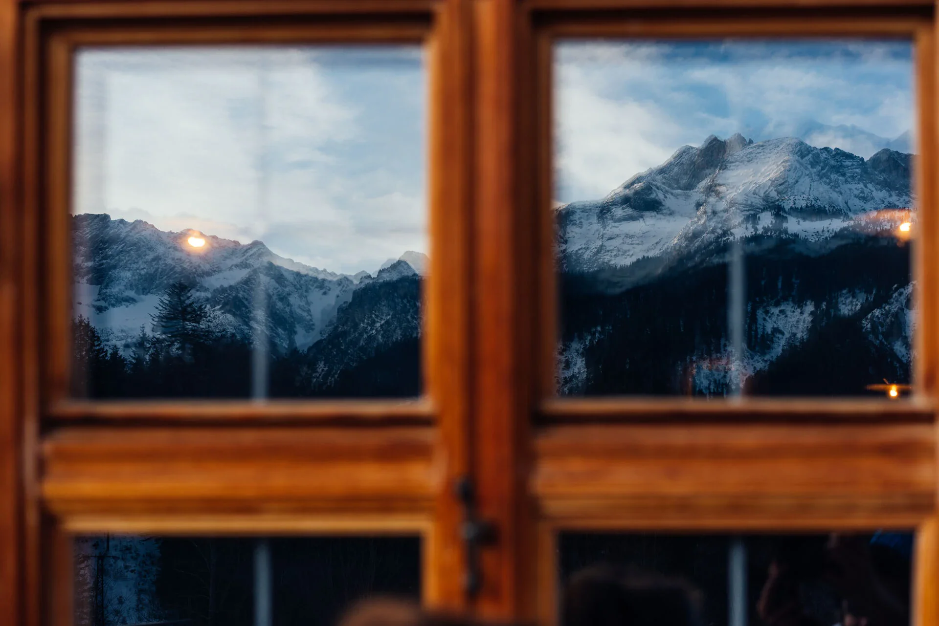 Durch ein Fenster mit braunem Rahmen hindurch ist eine schneebedeckte Berglandschaft bei tief stehender Sonne zu sehen.