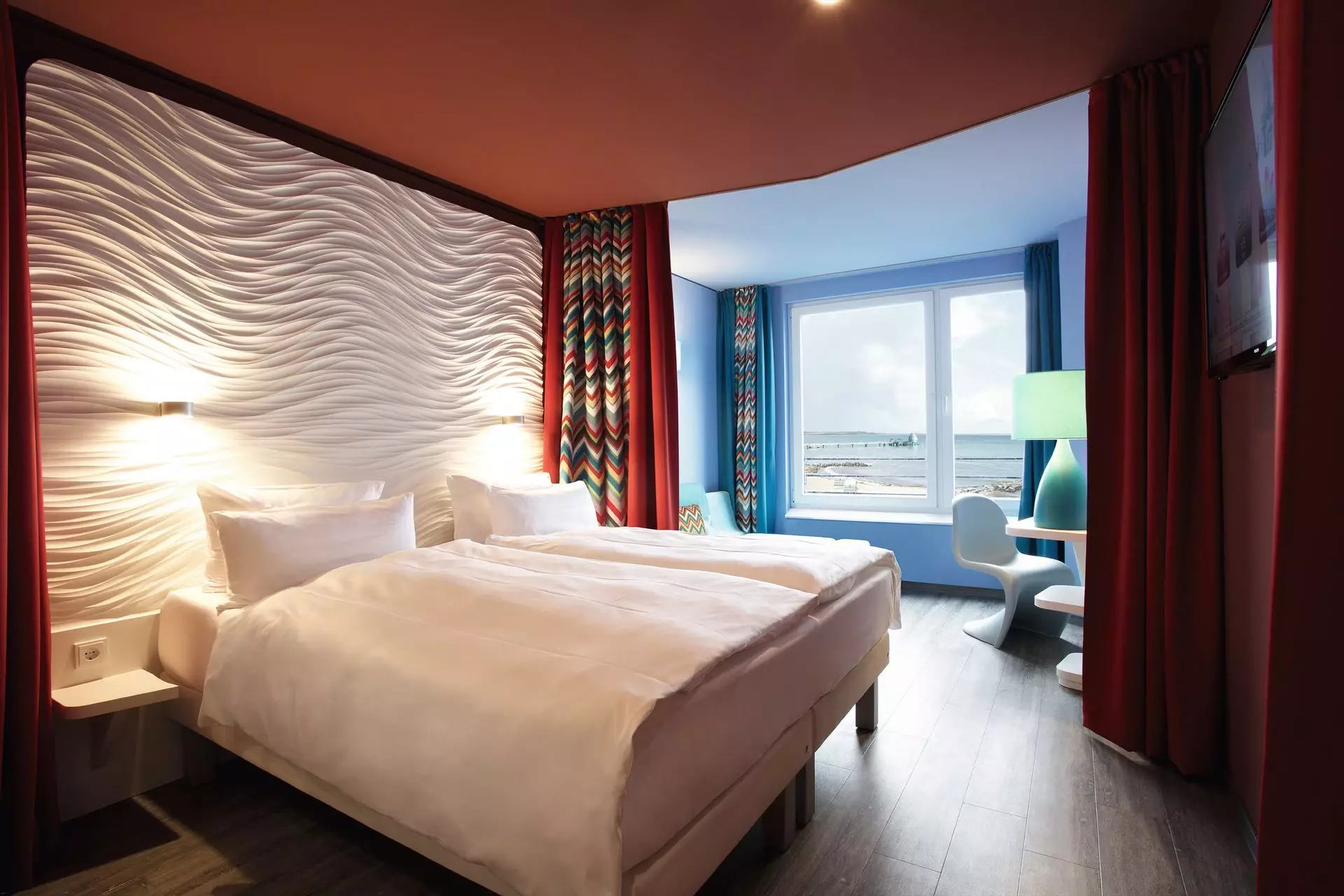Hotelzimmer im aja Grömitz Ein mit weißer Bettwäsche bezogenes Doppelbett steht vor einer weißen Rückwand mit Struktur. Umrahmt von einer dunkelroten Wand und einem rot-gemusterten Vorhang steht das Bett in einem Raum mit rostroter Decke und dunklem Holzboden. Im Hintergrund ist ein von blauen Wänden umrahmtes Fenster mit Blick auf die Ostsee zu erkennen.