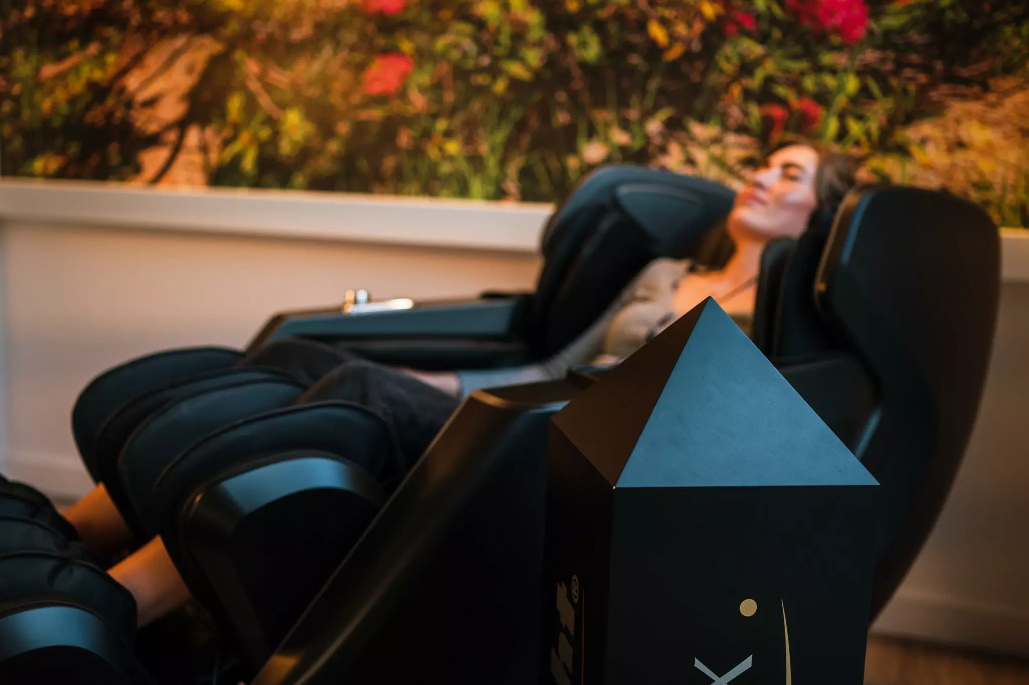 Massage-Sessel Eine Frau sitzt entspannt und lächelnd in einem schwarzen Massagesessel vor einer Wand mit einem großen Bild von einer Naturaufnahme.
