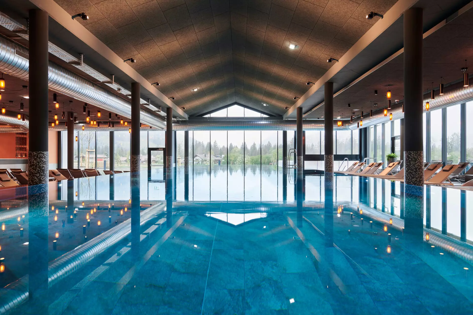Ein großer Innenpool der auf eine Glasfront zuläuft. Um den Pool stehen Liegen und kleine warme Lampen an der Decke spiegeln sich auf dem Wasser.