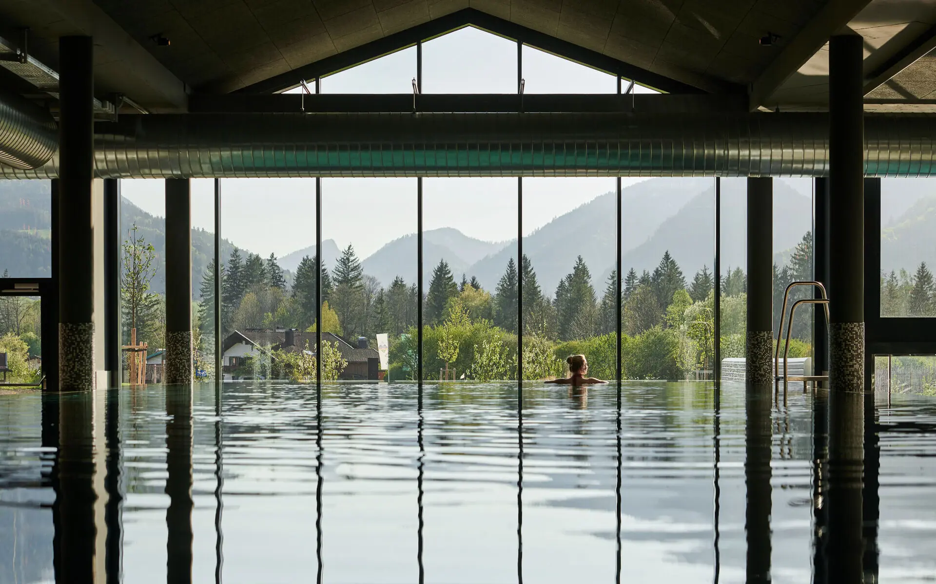Innenpool aja Ruhpolding Blick über den Innenpool des aja Ruhpolding. Im Hintergrund ist über dies gesamte Breite ein Panoramafenster mit Ausblick auf die grünen Bäume vor einer Bergkulisse dahinter zu sehen. Eine Frau stützt sich mit Blick nach draußen hinten auf den Beckenrand.