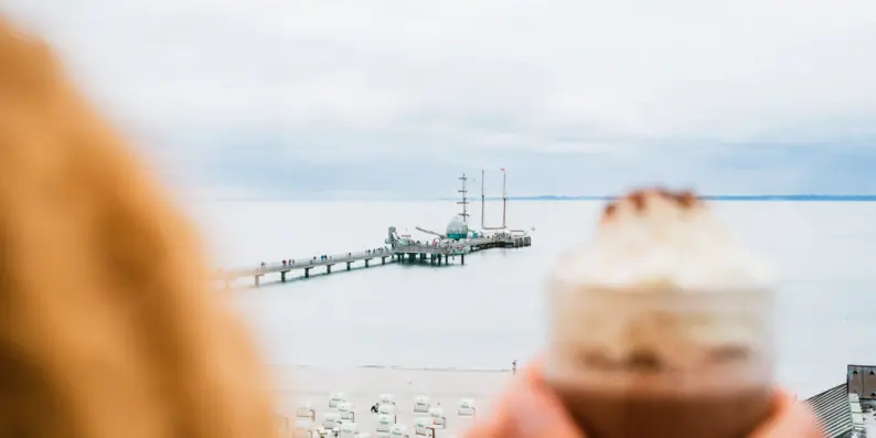Eine Frau steht mit ihrer heißen Schokolade an einem Fenster und schaut auf das graue Meer und den Steg. Eine Frau steht mit ihrer heißen Schokolade an einem Fenster und schaut auf das graue Meer und den Steg.