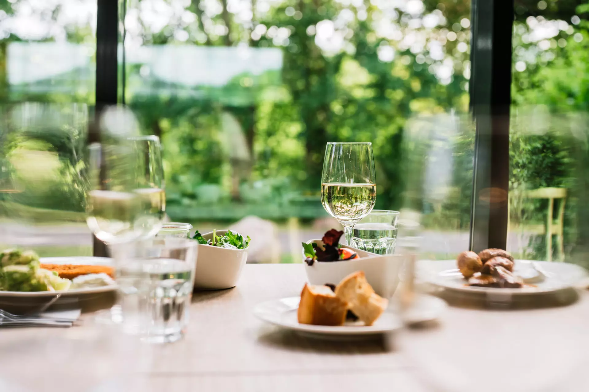Abendessen im aja Bad Saarow Ein Tisch ist mit Abendessen, Salatschälchen und Weingläsern gedeckt. Im Hintergrund gibt eine große Fensterfront den Blick auf die dahinter liegende Natur frei.