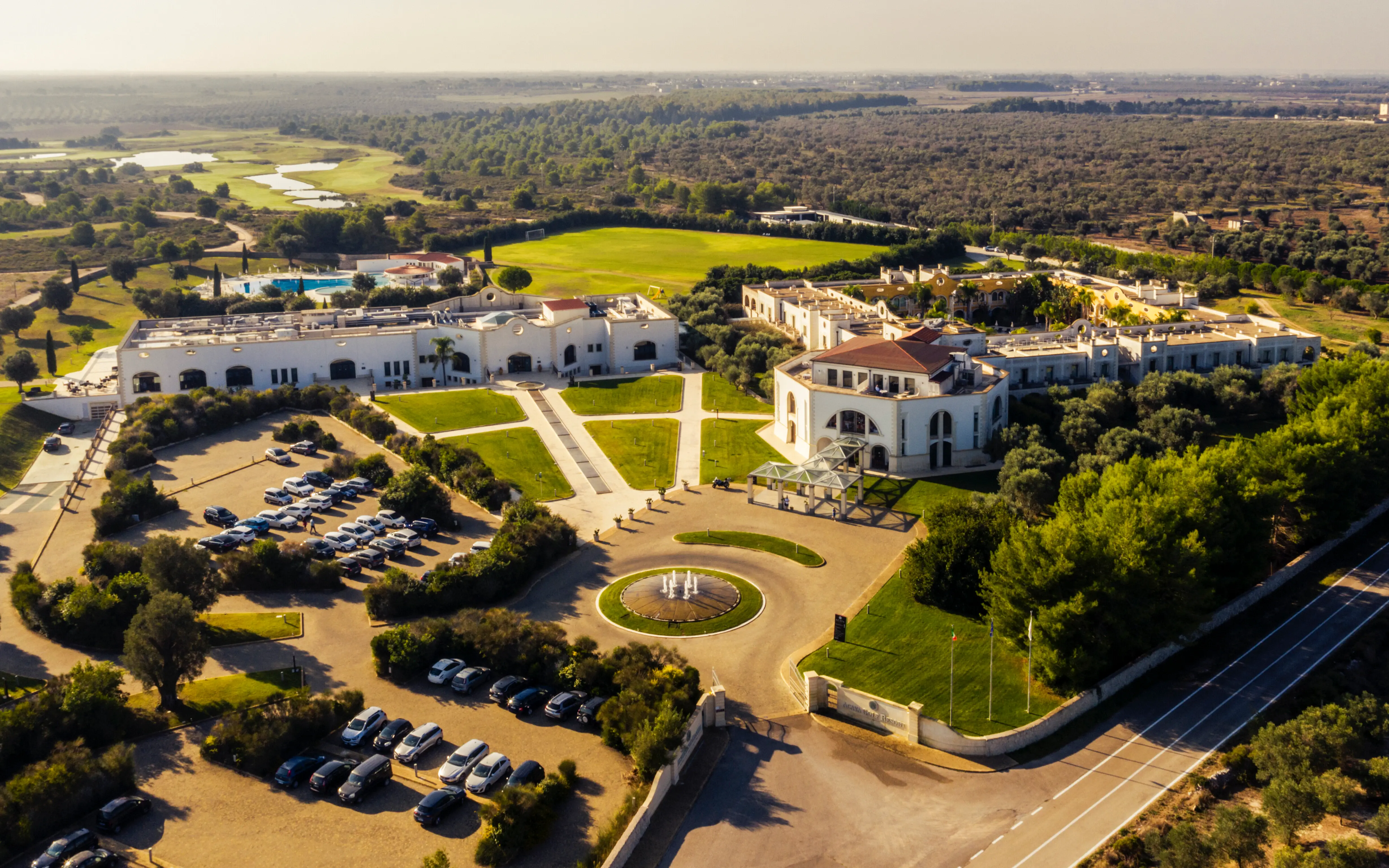 Acaya Golf Resort & Spa Luftaufnahme des Acaya Golf Resort & Spa, eingebettet in die mediterrane Landschaft Süditaliens, mit gepflegten Golfplätzen, eleganten weißen Gebäuden, einem zentralen Springbrunnen und umgeben von Olivenhainen und Natur.