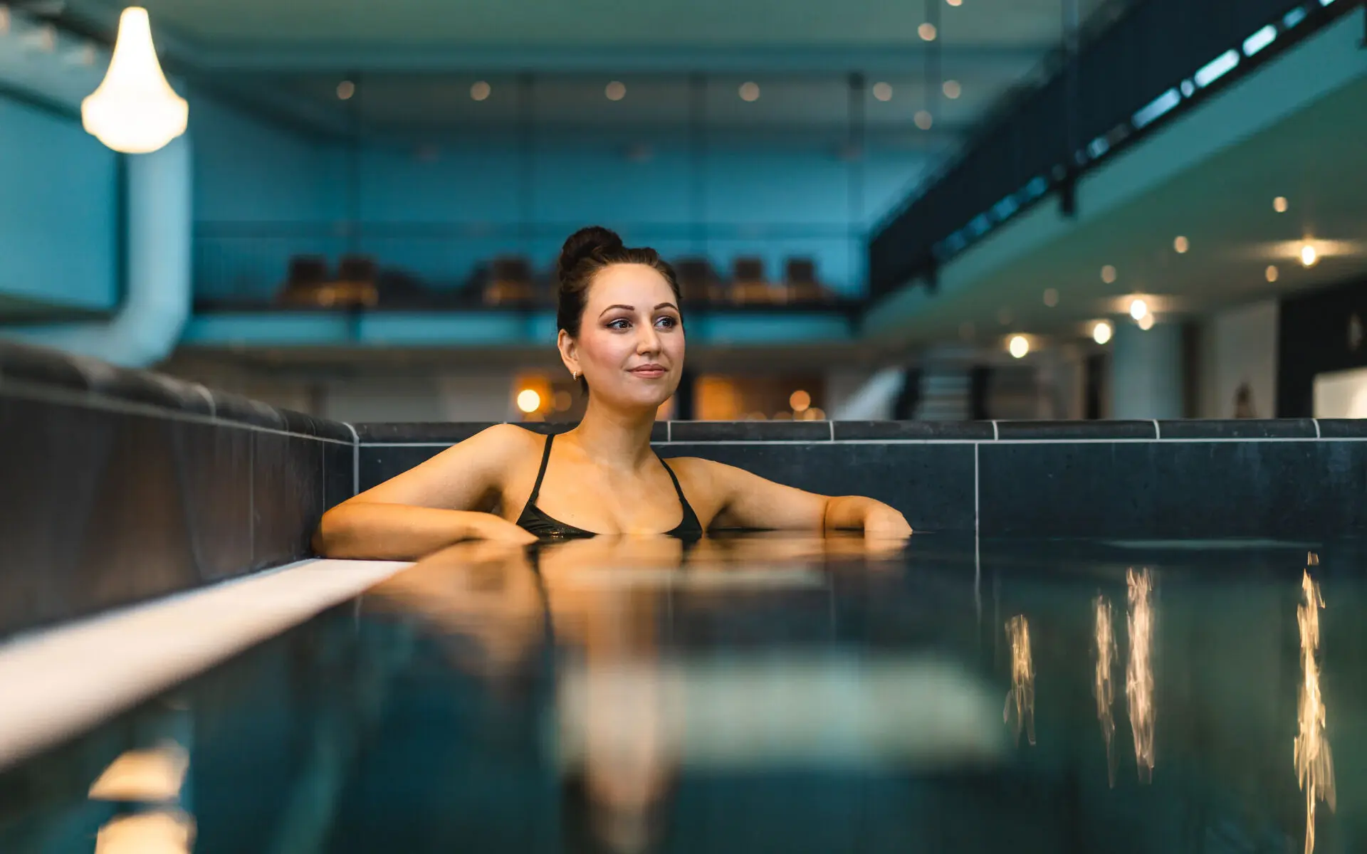 Whirlpool aja Travemünde Eine dunkelhaarige Frau entspannt in einem eckigen Schwimmbecken, die Ellenbogen auf dem Beckenrand aufgestützt. Im Hintergrund ist abgedunkelt und leicht verschwommen der Schwimmbereich zu erkennen.