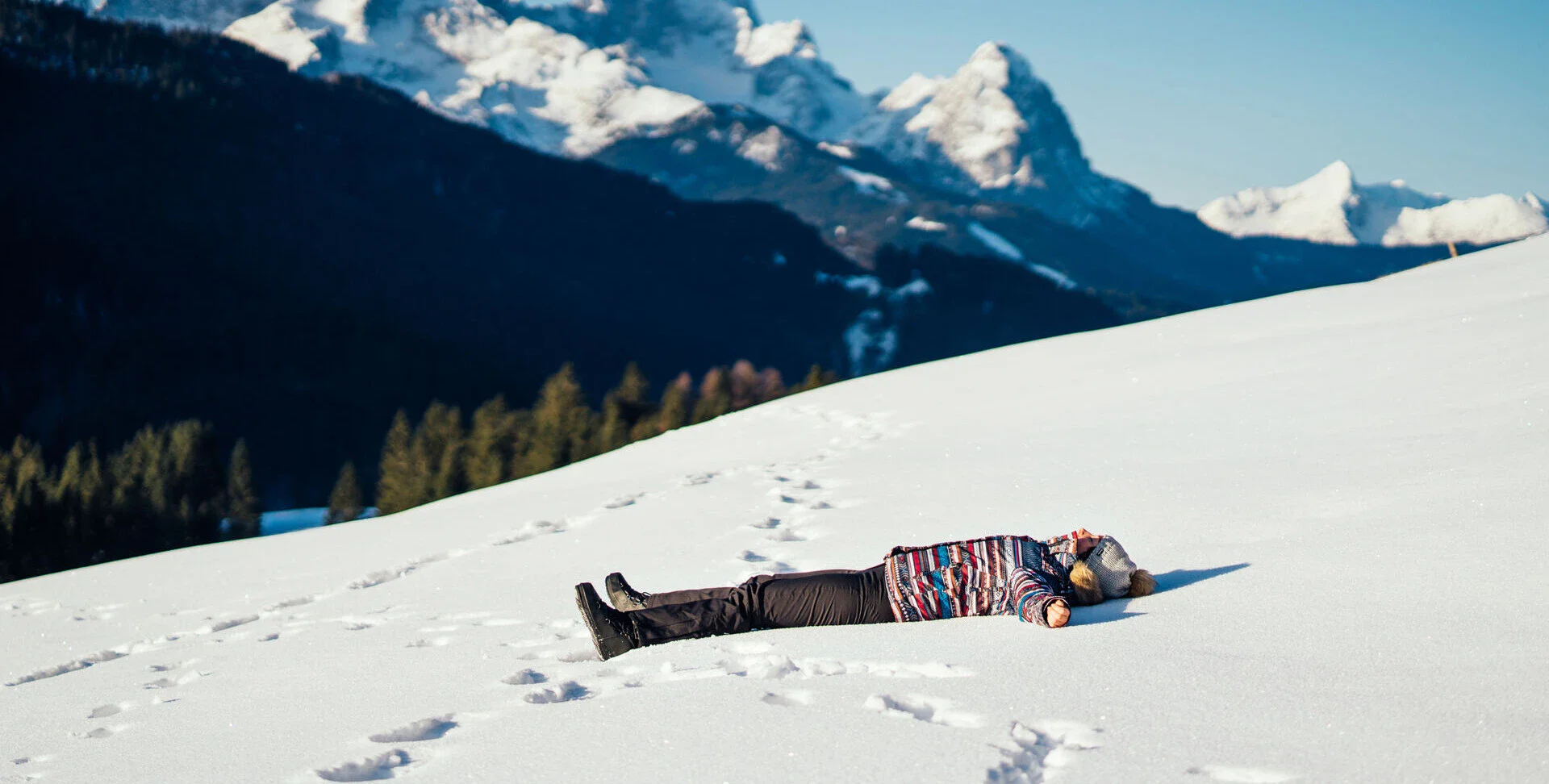Winterurlaub Eine Frau in Winterkleidung liegt an einem Hang im Schnee. Die Sonne scheint und im Hintergrund erstreckt sich eine Berg- und Waldlandschaft.