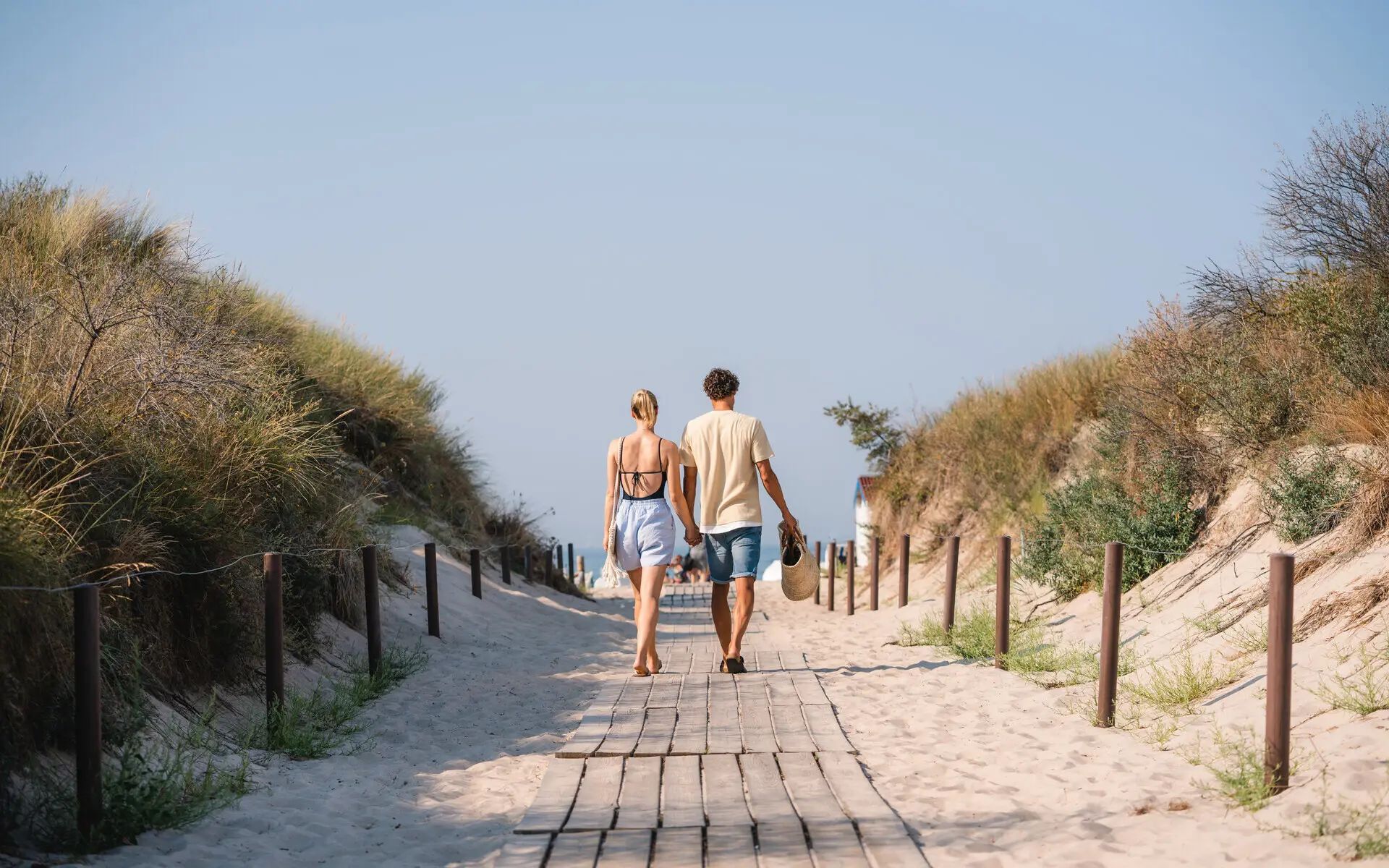 Warnemünde Ein Paar läuft Hand in Hand einen Weg zwischen Denen zum Strand hinunter. Die Sonne scheint warm und der Himmel ist blau.