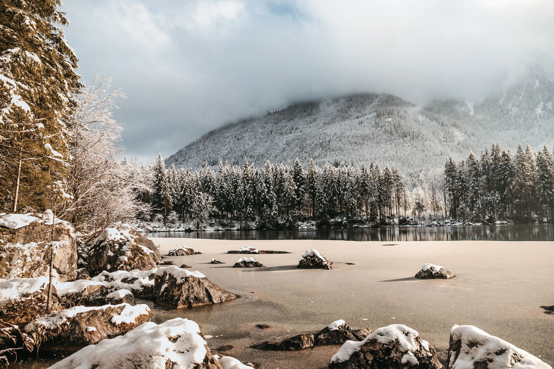 Verschneiter See mit schneebedeckten Felsen und umrahmt von dichten Tannenwäldern, vor einer beeindruckenden Bergkulisse, die teils in Nebel gehüllt ist.
