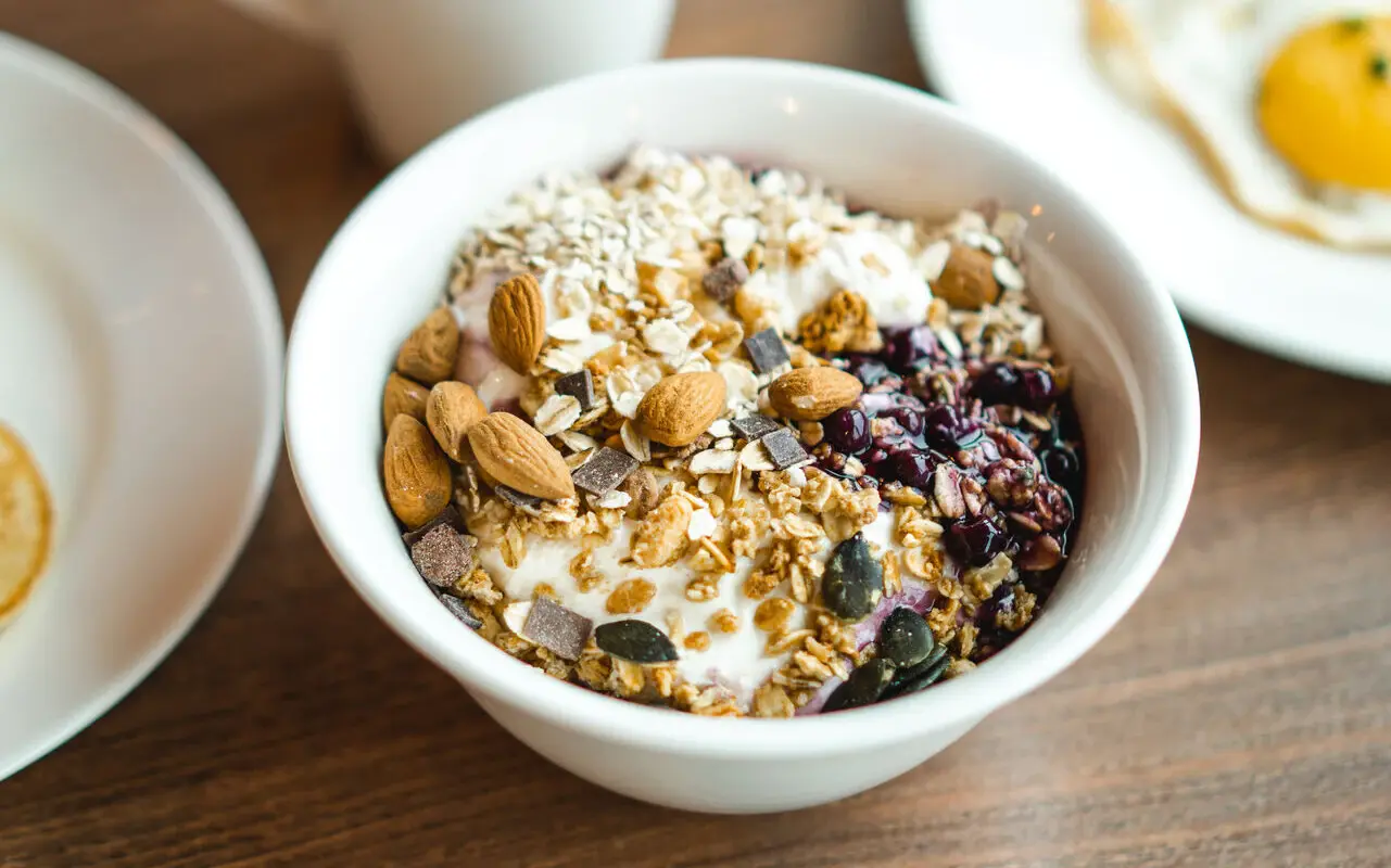Müsli Auf einem Holztisch steht eine Müslischale aus Jogurt, Haferflocken, Nüssen und getrockneten Beeren. Daneben sind die Ränder weiterer Teller zu sehen.