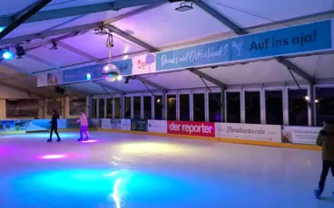Verschiedenen Personen fahren auf einer Eisbahn Schlittschuh. Bunte Farben leuchten von der Discokugel auf das Eis.