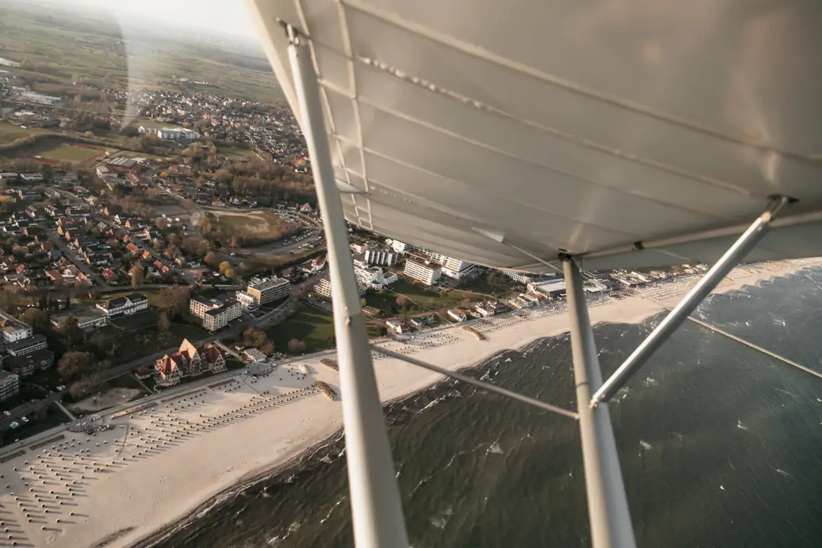Flug über Grömitz Flugzeugflügel über einem Strand mit Gebäuden im Hintergrund.