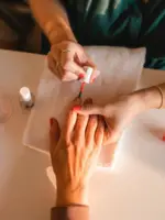 Von oben fotografiert werden die Nägel einer Frau rot lackiert. Die Kosmetikerin hat Ringe an ihren Fingern und setzt mit dem Pinsel zum Lackieren des Zeigefingers an.