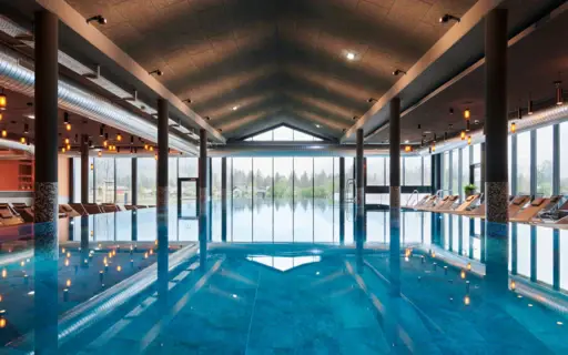 Innenpool aja Ruhpolding mit ruhigem Wasser, auf dem sich kleine Lampenlichter Spiegeln, und einer großen Fensterfront im Hintergrund. Innenpool aja Ruhpolding mit ruhigem Wasser, auf dem sich kleine Lampenlichter Spiegeln, und einer großen Fensterfront im Hintergrund.
