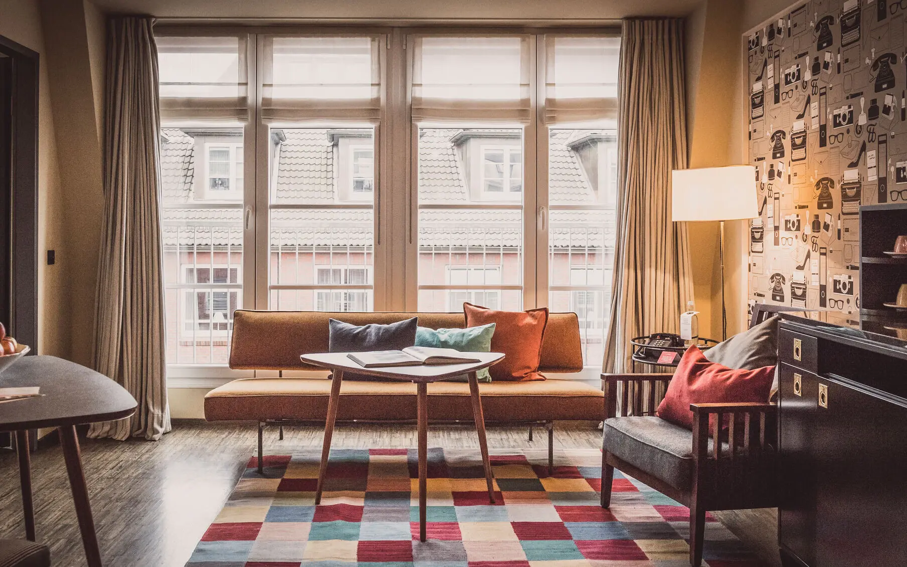 HENRI Hamburg Gemütliches Wohnzimmer im HENRI Hamburg mit einer Retro-Couch, bunten Kissen, einem modernen Teppich, einem eleganten Holztisch und einer großen Fensterfront mit Blick auf die umliegenden Gebäude.