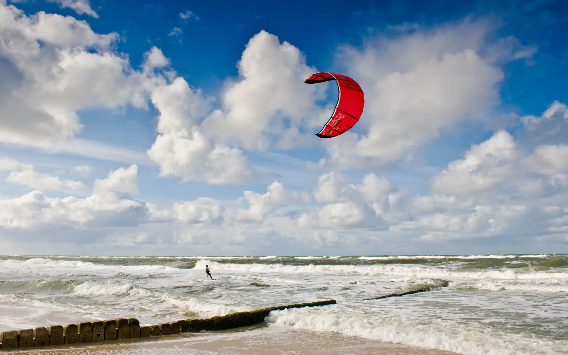 Auf welligem Wasser fährt ein Kitesurfer mit einem roten Kite an einem sonnigen Tag.
