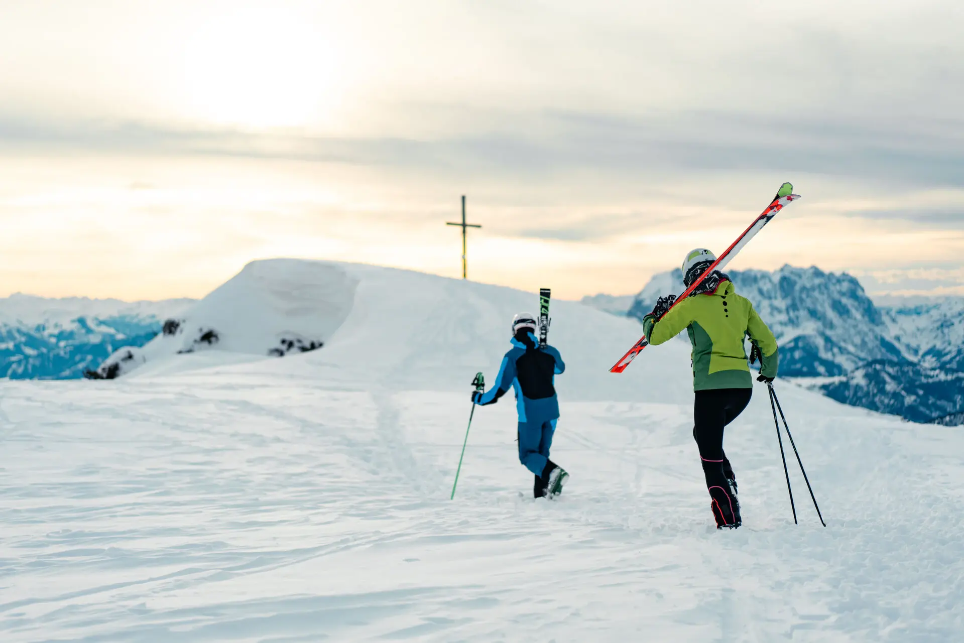 Zwei Skifahrer wandern mit ihren Skiern über eine schneebedeckte Berglandschaft in Richtung eines Gipfelkreuzes. Im Hintergrund sind beeindruckende Bergketten unter einem sanften, wolkenverhangenen Himmel zu sehen.