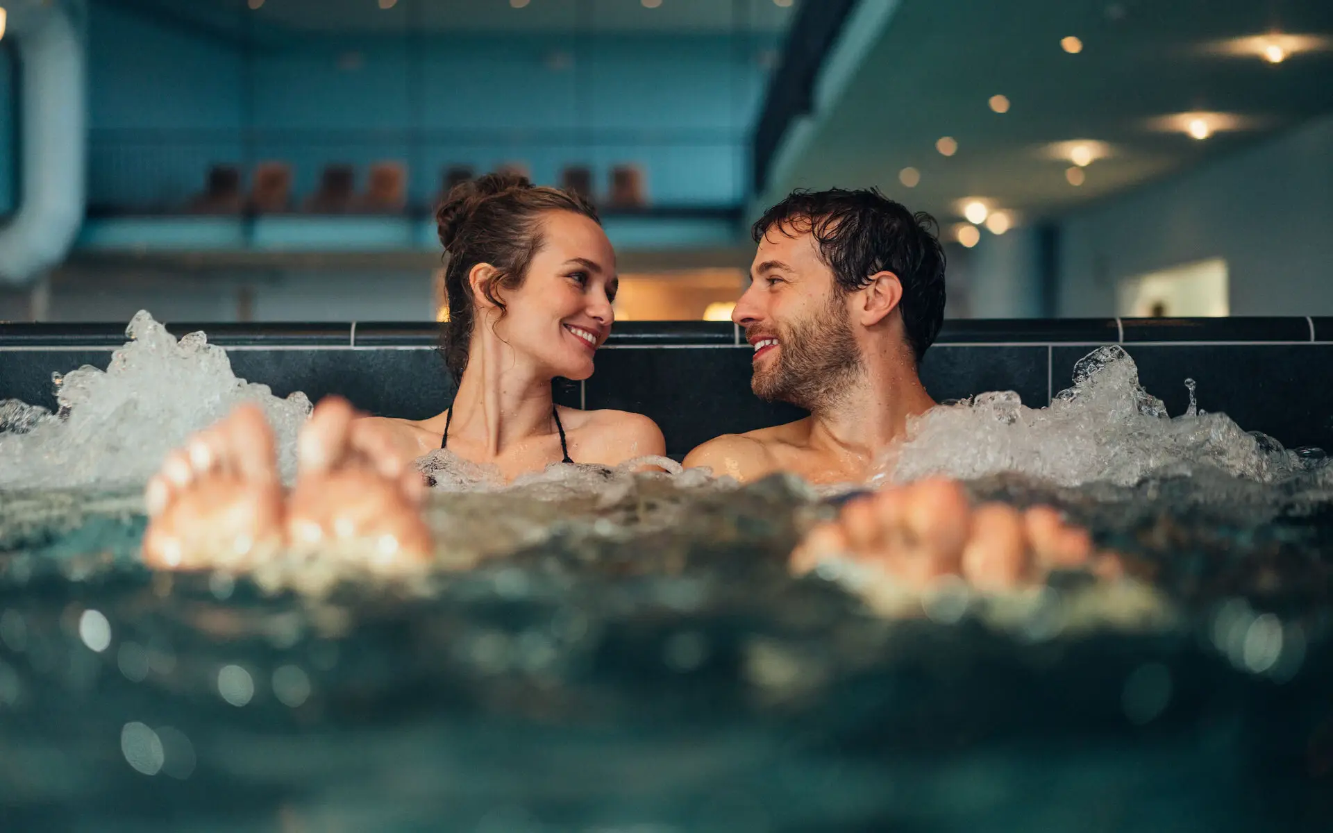 Whirlpool aja Warnemünde Ein Mann und eine Frau sitzen in einem sprudelnden Schwimmbecken, die Blicke einander zugewandt. Im Hintergrund ist abgedunkelt und leicht verschwommen einige Liegen im oberen Bereich zu erkennen.