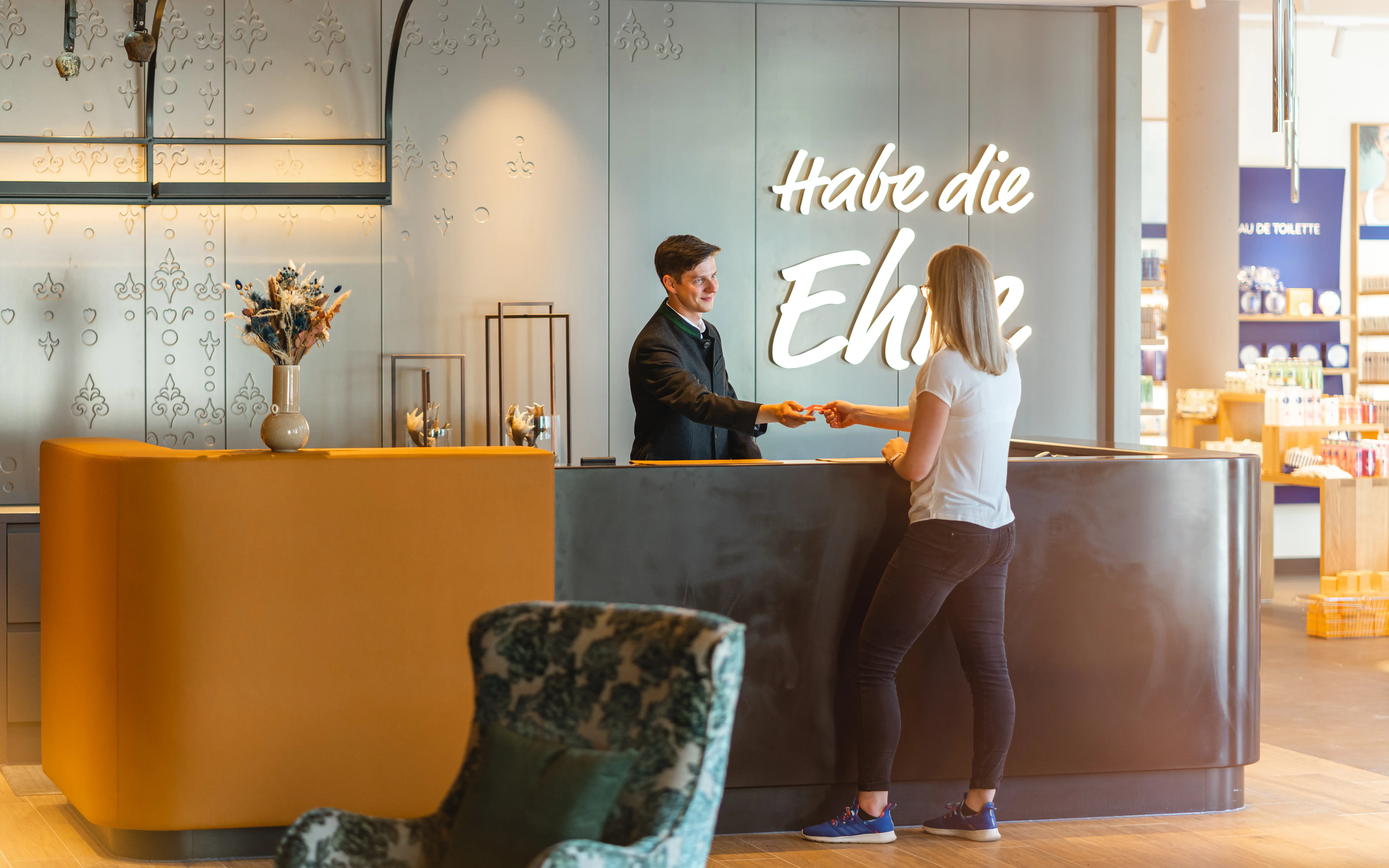 Empfangsbereich eines Hotels mit stilvollem Design. Ein freundlicher Rezeptionist überreicht einer Gästin eine Schlüsselkarte. Im Hintergrund ist eine Wand mit dem Schriftzug „Habe die Ehre“ beleuchtet.