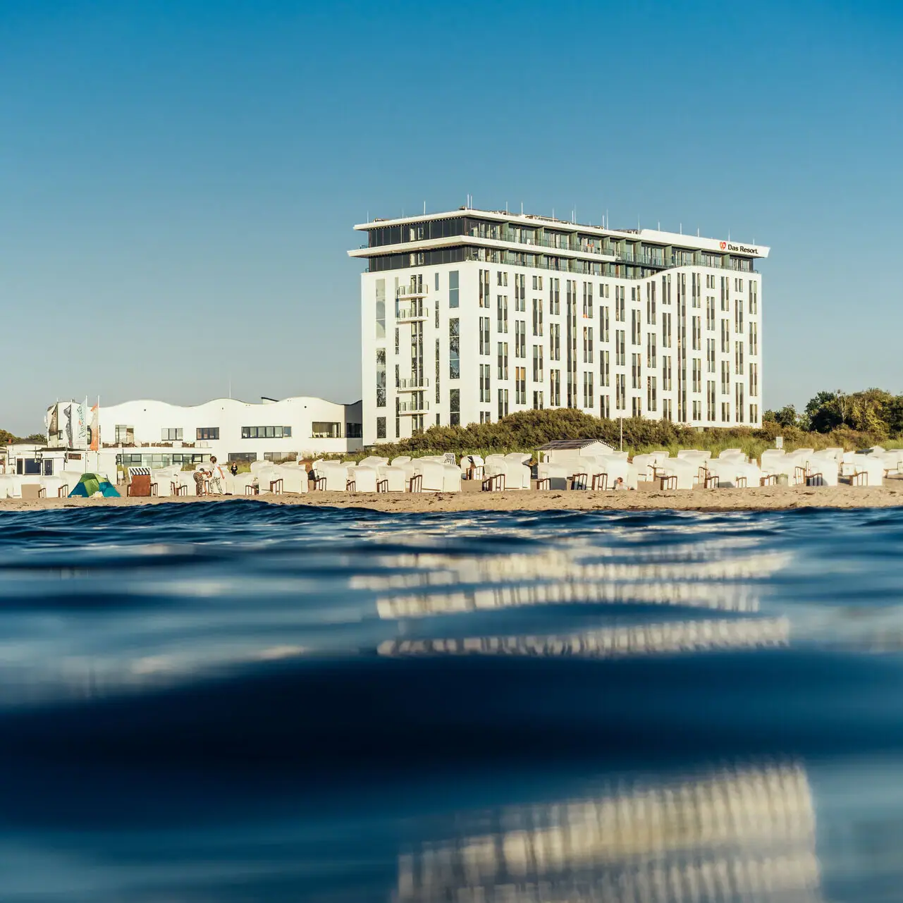 Das Hotelgebäude am Strand mit klarem Himmel und Wasser im Vordergrund.