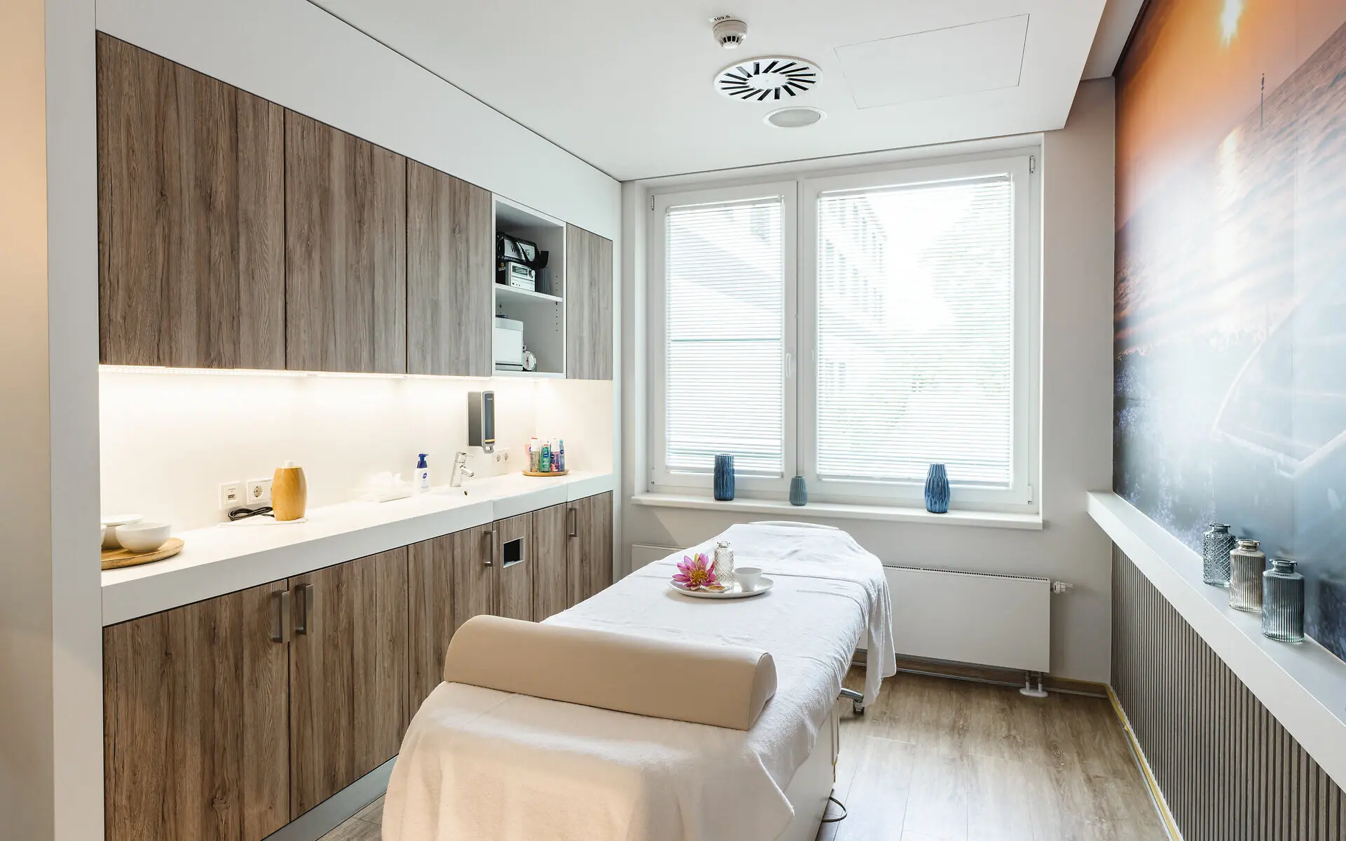 Anwendung im NIVEA Haus Bad Saarow Ein Anwendungsraum in hellen Farben, mit einer Behandlungsliege, holzverkleideten Schränken und einem großen Fenster, durch welches Tageslicht in den Raum scheint. An der rechen Wand hängt ein großes Bild der Ostsee bei Sonnenuntergang.