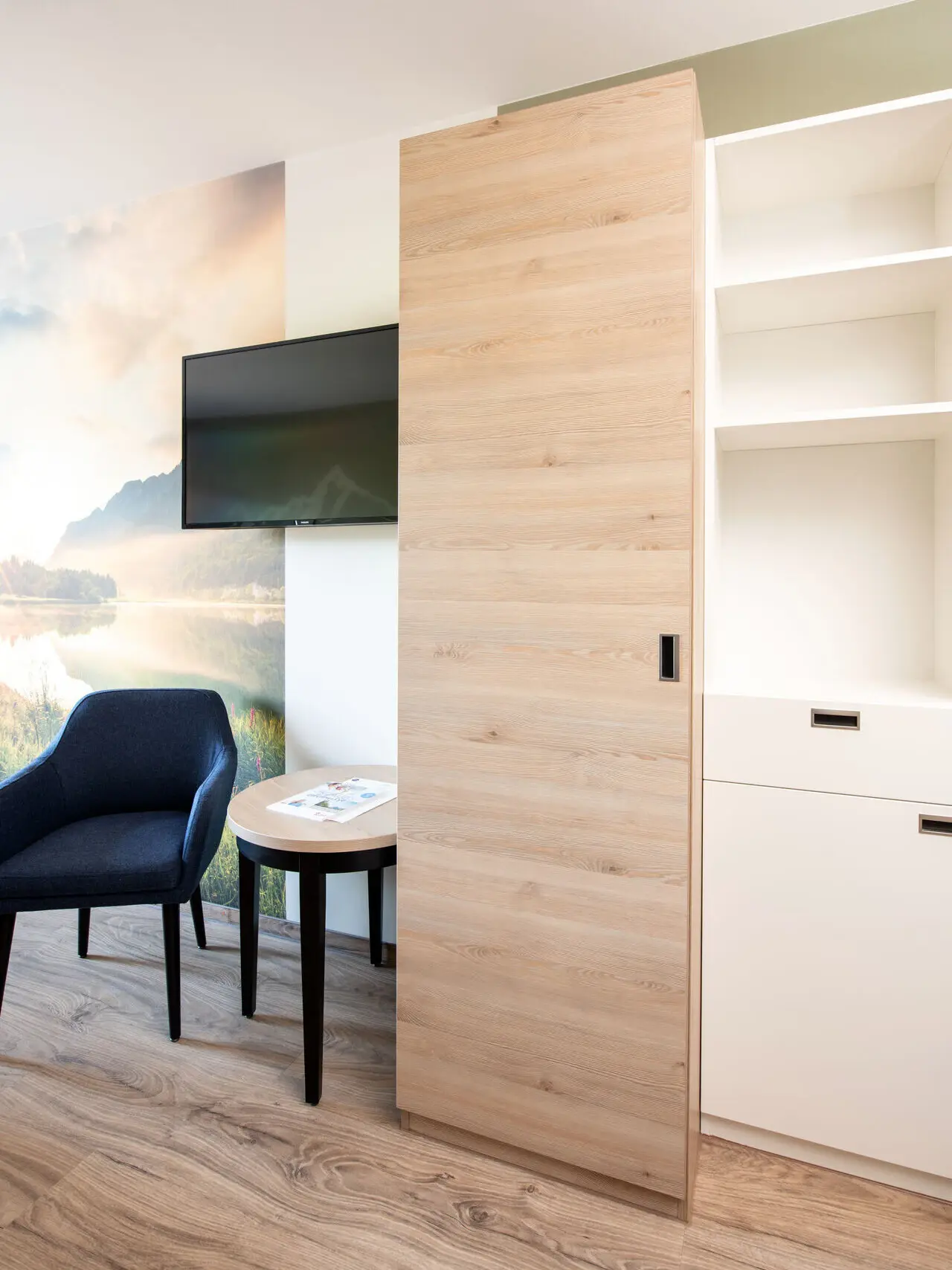 Ein Ausschnitt von einem Hotelzimmer mit Holzboden und weißen Wänden. Neben einem Schrank mit Holztüren seht ein dunkler Stuhl mit einem kleinen Beistelltisch und darüber hängt ein Fernseher.