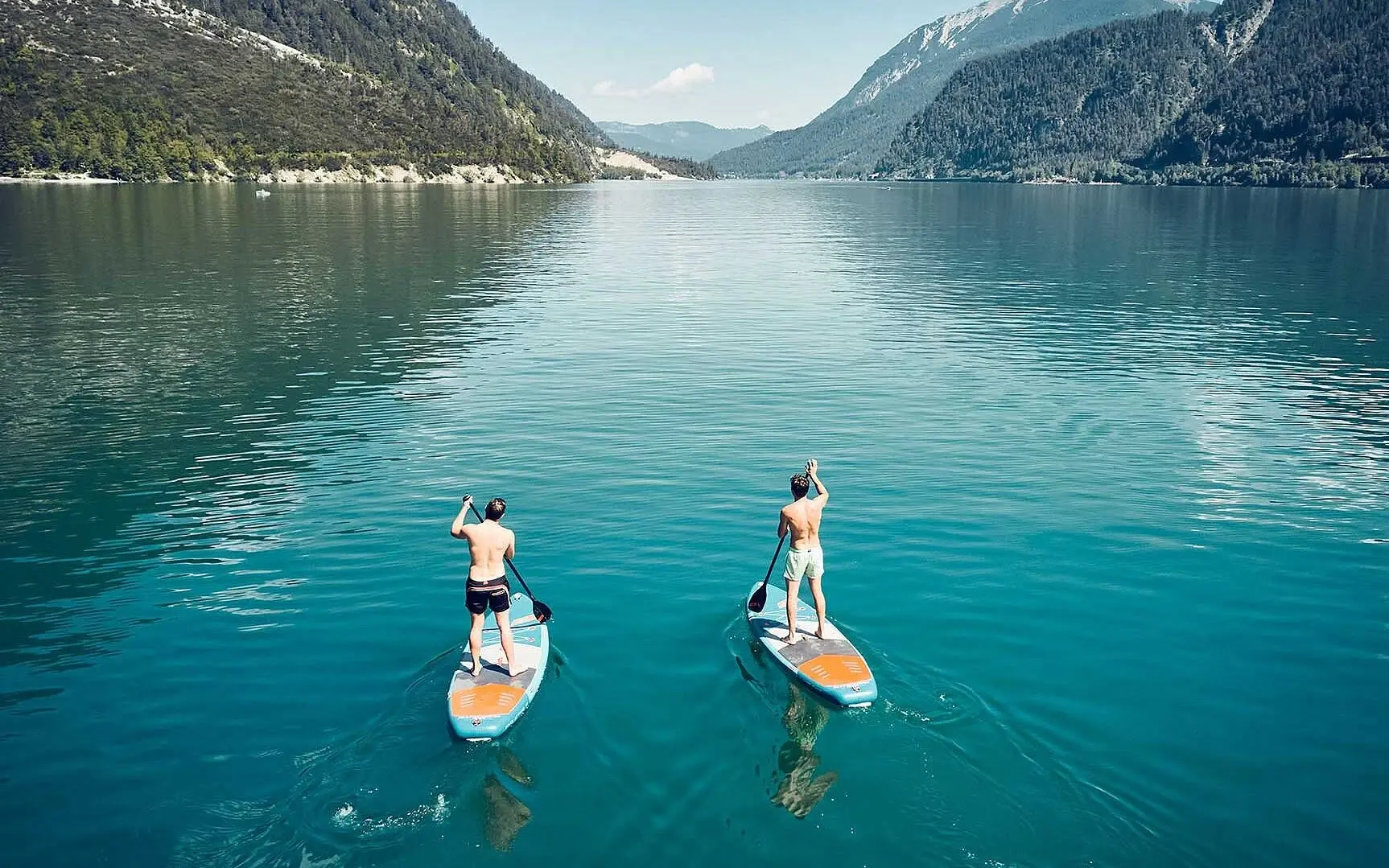 Zwei Männer auf Paddleboards auf einem See mit Bergen im Hintergrund.