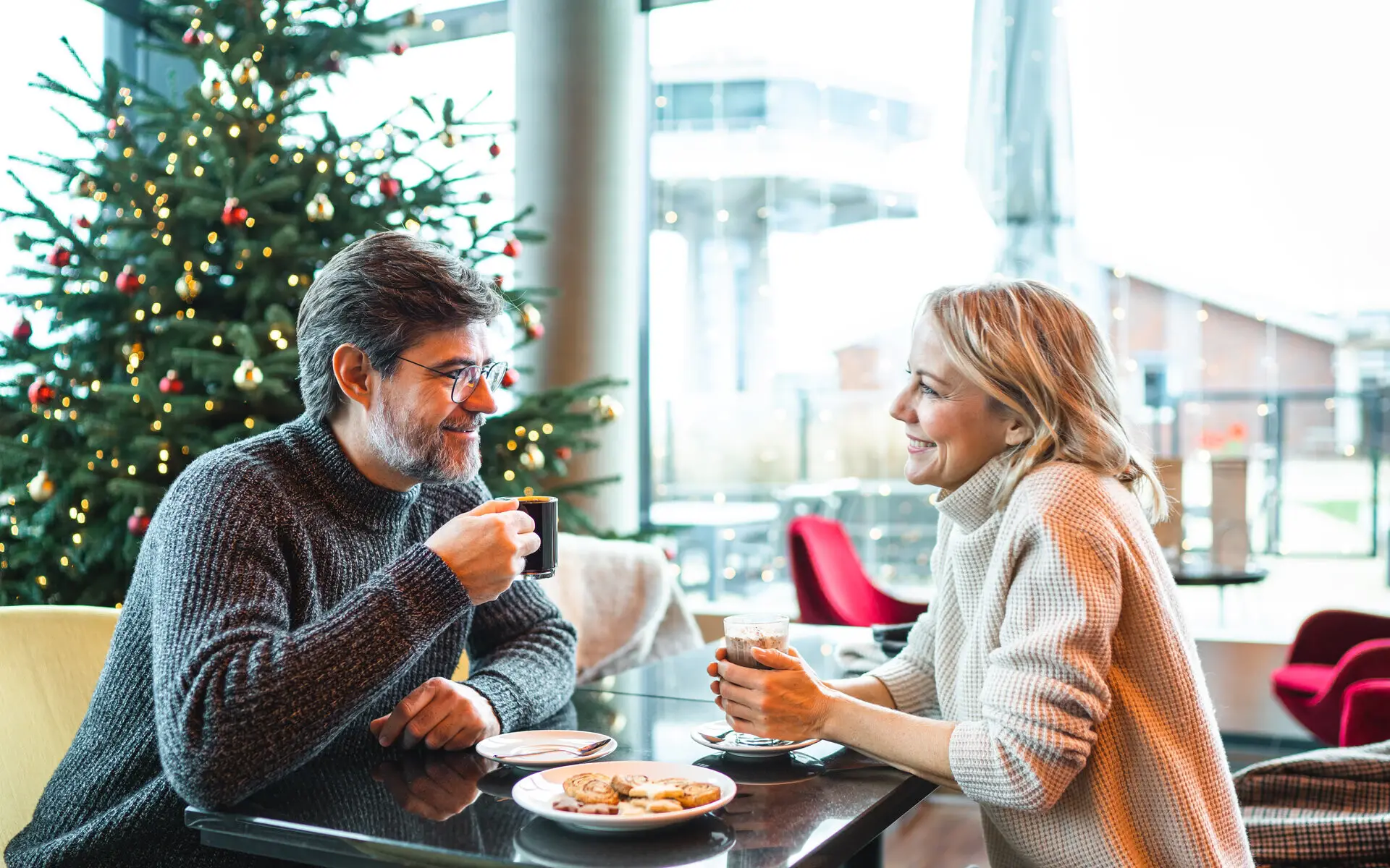 Latte Macchiato Ein lächelndes Paar sitzt in einer gemütlichen Lounge vor einem festlich geschmückten Weihnachtsbaum. Beide genießen warme Getränke und unterhalten sich, während Kekse auf dem Tisch serviert sind.