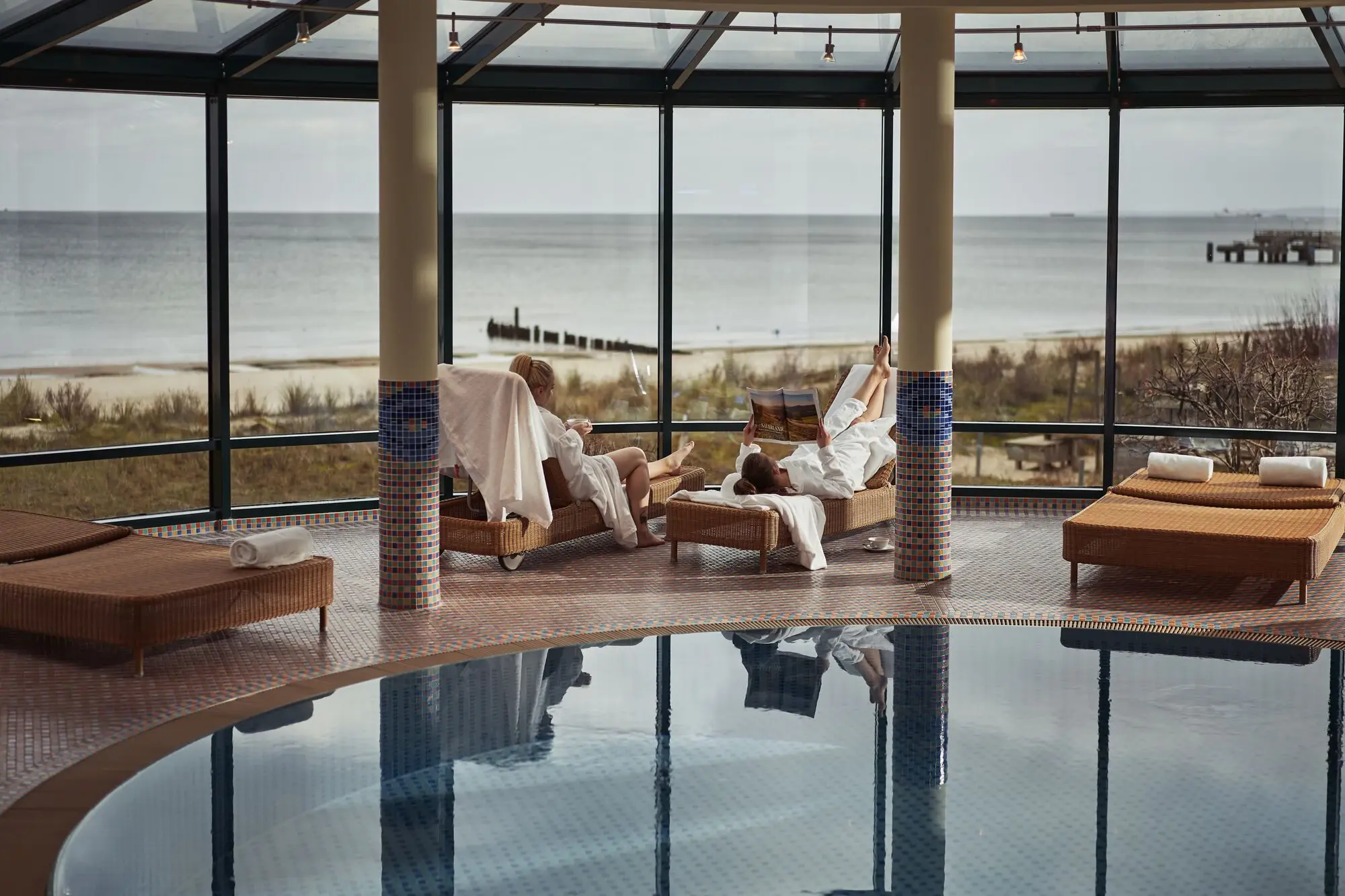 aja Strandhotel Bansin Zwei Personen in Bademänteln entspannen auf Liegen in einem lichtdurchfluteten Spa-Bereich mit Panoramablick auf den Strand und die Ostsee.