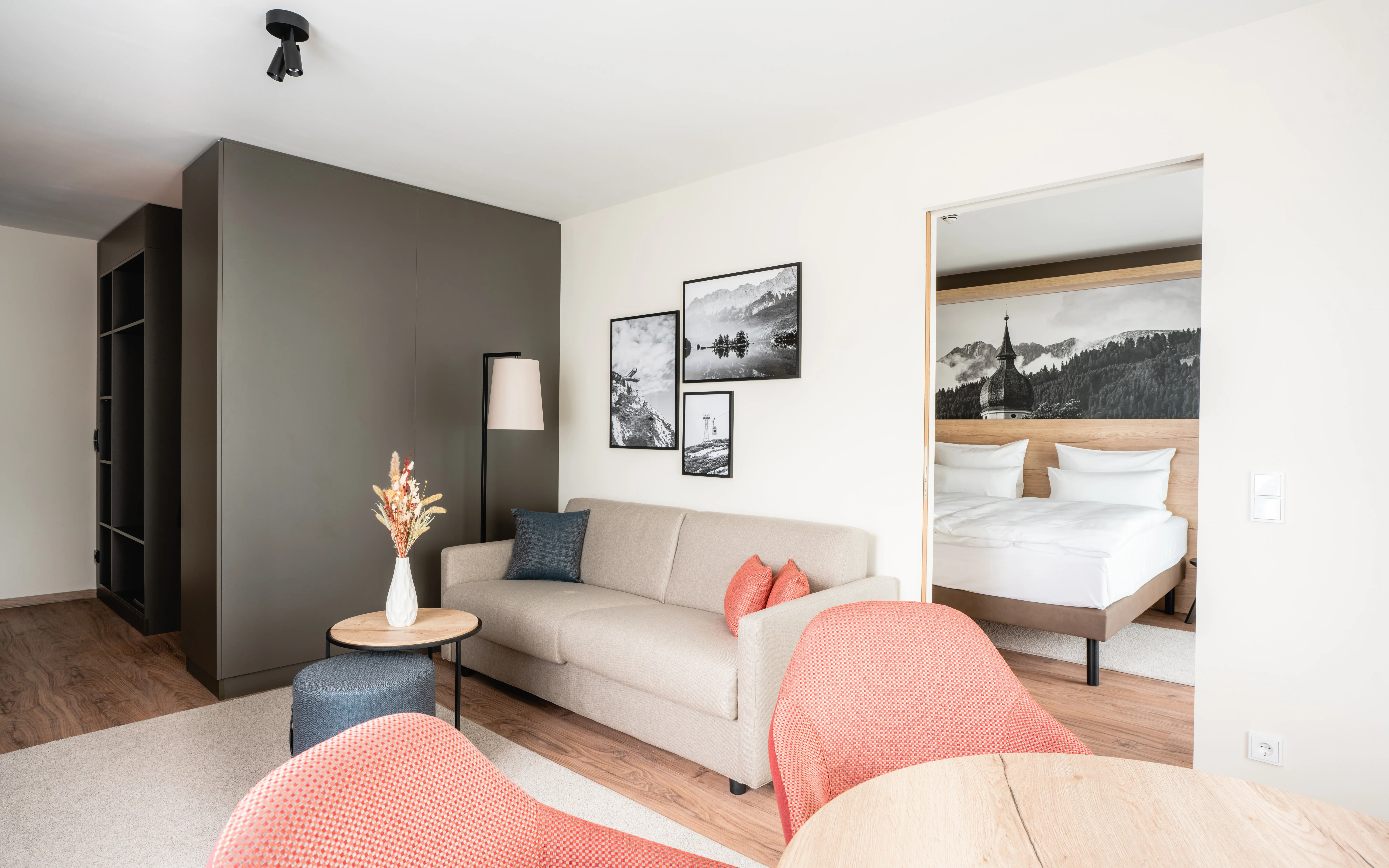 Helles und modernes Hotelzimmer mit separatem Wohn- und Schlafbereich. Der Wohnbereich ist mit einem Sofa, einem Sessel und einem kleinen Tisch mit Trockenblumen dekoriert. An der Wand hängen schwarz-weiße Landschaftsbilder.Im Hintergrund ist der Schlafbereich mit einem großen Doppelbett und einer Wanddekoration mit Bergmotiv zu sehen.