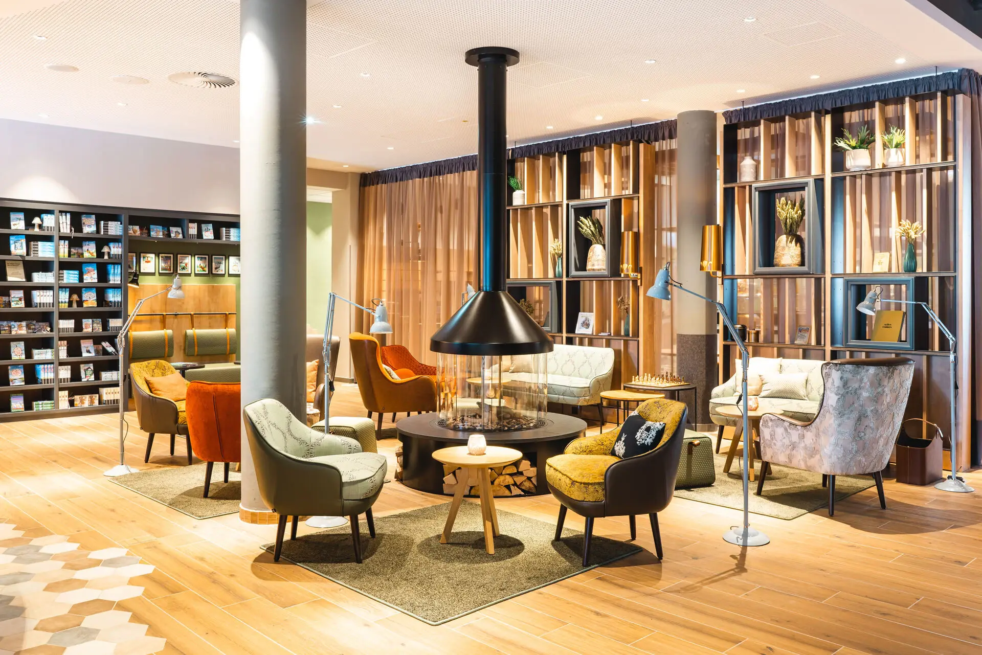 Gemütlicher Lounge-Bereich mit einem zentralen, modernen Kamin, umgeben von bequemen Sesseln und Sofas in verschiedenen Farben und Stoffen. Im Hintergrund ist eine Bücherauswahl zu sehen, die zum Stöbern einlädt.