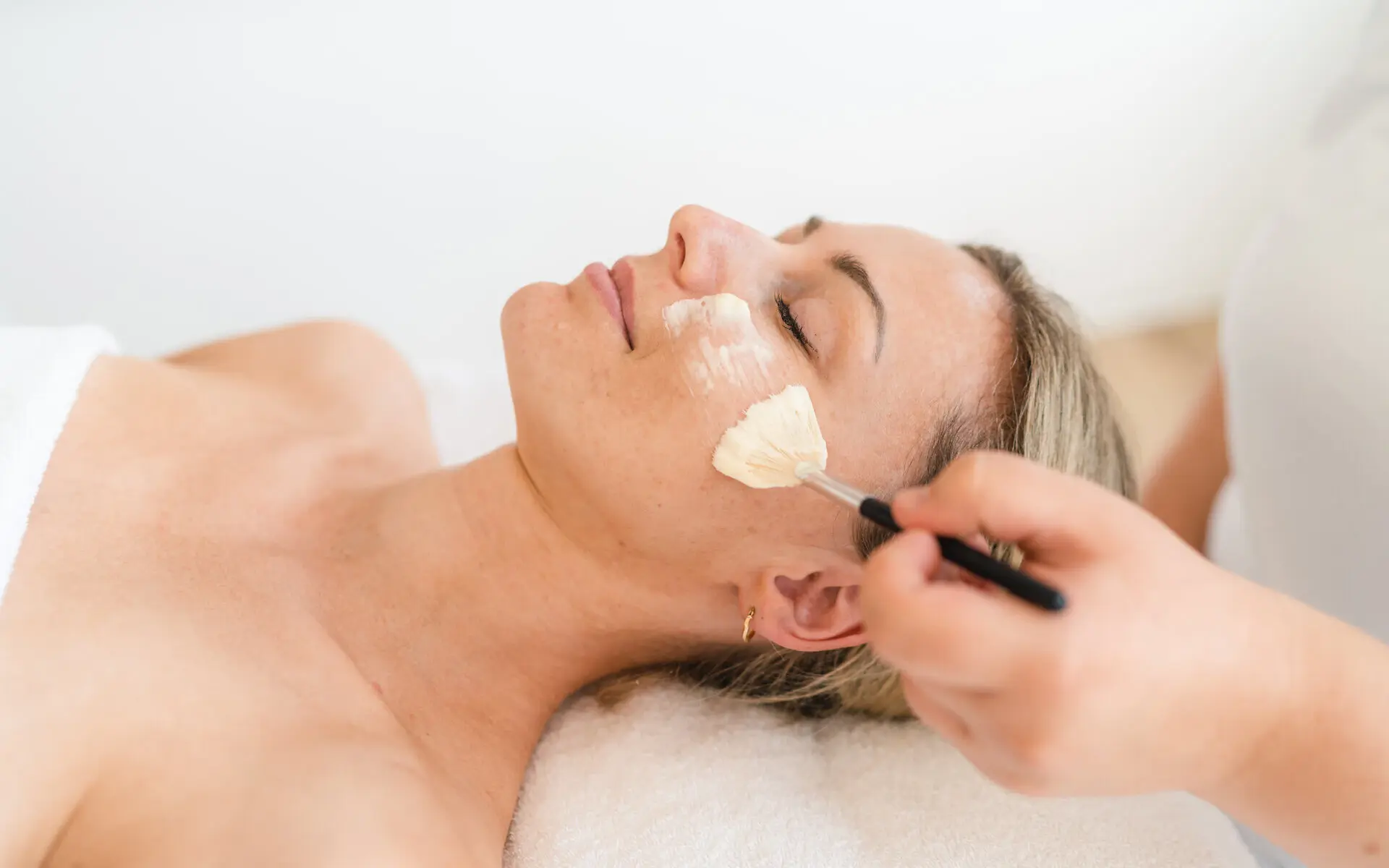 Kosmetik im NIVEA Haus Eine Frau liegt auf dem Rücken und erhält eine Gesichtsanwendung mit einem Kosmetikpinsel. Der Kopf der Frau liegt entspannt ab, das Gesicht ist zur Decke gerichtet. Die Augen der Frau sind geschlossen, sie lächelt leicht und ihre Gesichtszüge wirken entspannt.