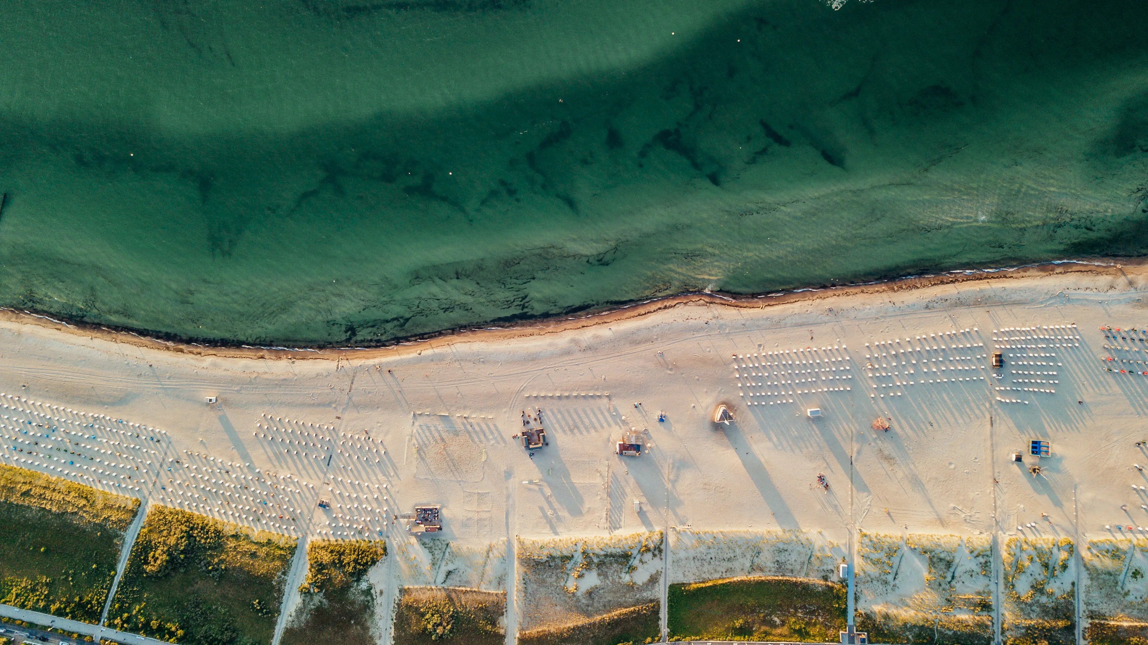 Luftaufnahme eines breiten Sandstrandes mit geordneten Sonnenliegen, angrenzender Vegetation und dem ruhigen, grünen Meer im Hintergrund.