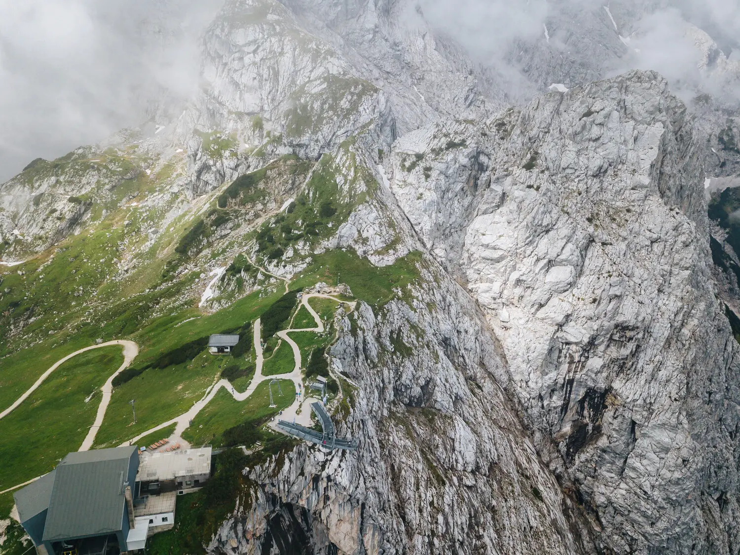 Eine beeindruckende Luftaufnahme von einer Berglandschaft mit steilen Felswänden und grünen Wiesen. Ein gewundener Wanderweg führt zu einer Aussichtsplattform und einer Berghütte.