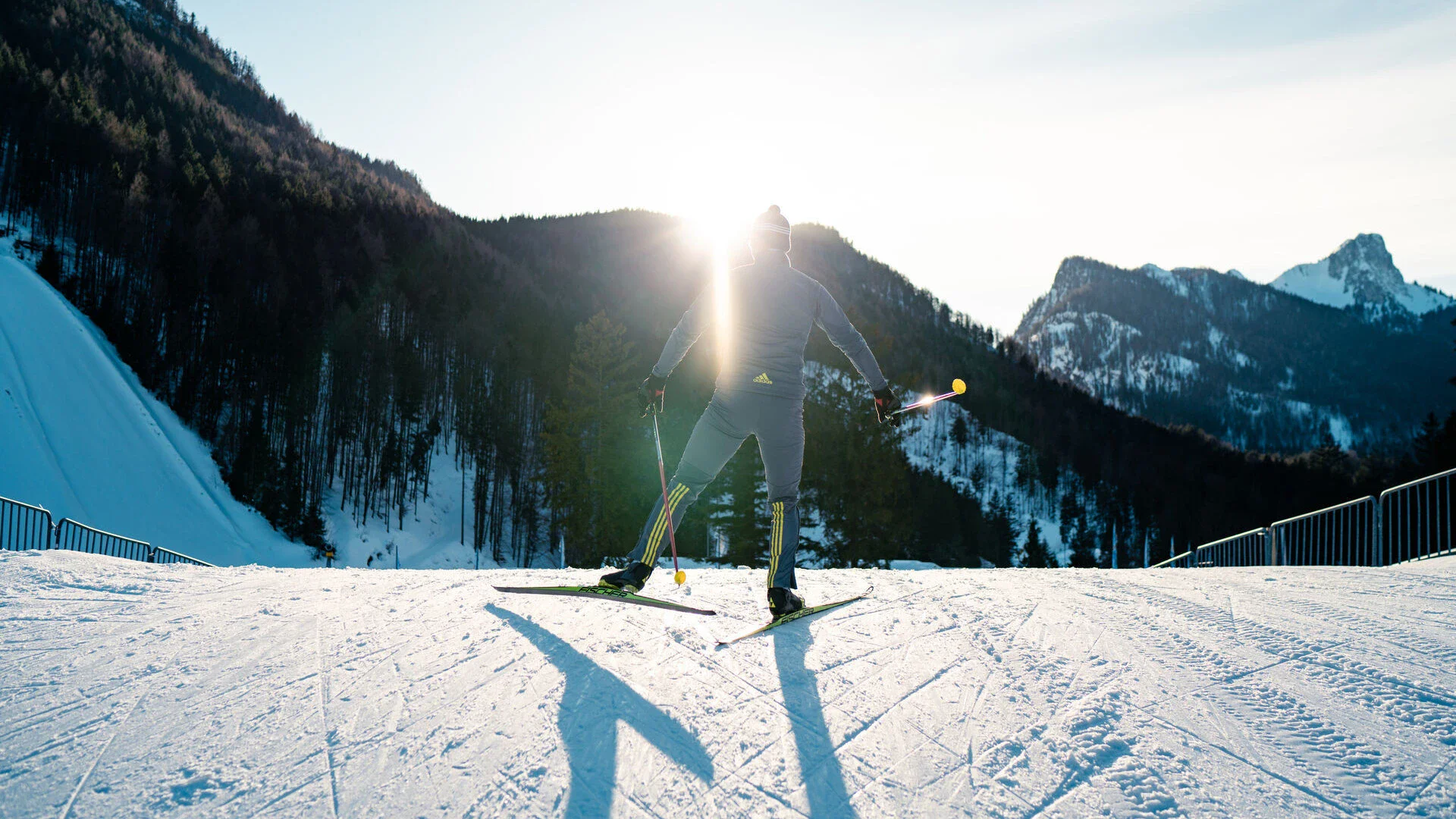 Ein Mann auf Skiern fährt eine schneebedeckte Piste hinter. Im Hintergrund erstrecken sich mit Wäldern bewachsene Berge und die Sonne scheint am Himmel empor.