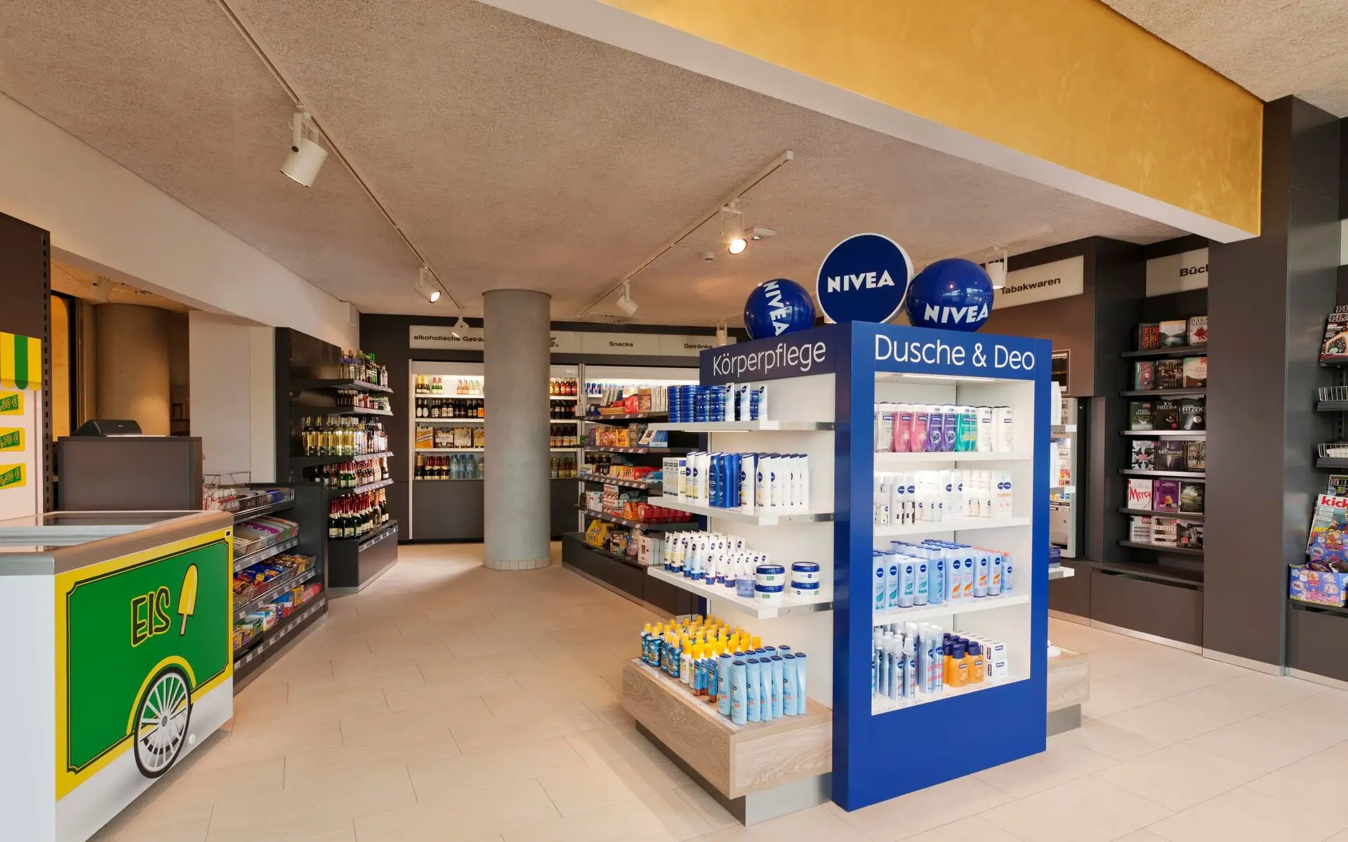 NIVEA Haus In einem Verkaufsraum mit hellem Holzboden und weißen Wänden sind diverse NIVEA Produkte in den Regalen zu erkennen. An der Decke im Hintergrund hängen blaue und weiße Luftballons mit dem NIVEA Logo bedruckt und links im Vordergrund ist ein weißer Verkaufstresen zu sehen.