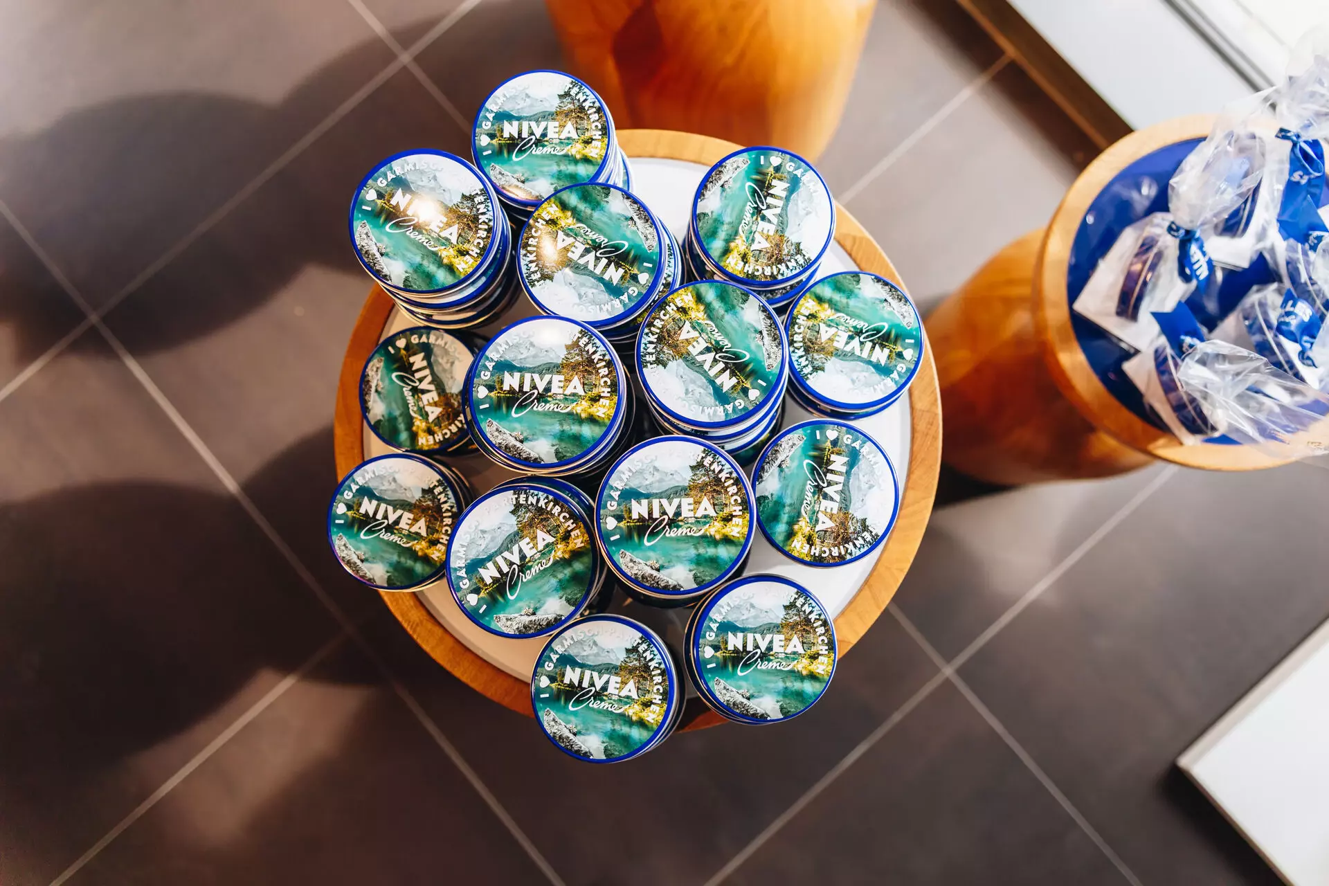 Runde Holzaufsteller mit mehreren Dosen NIVEA-Creme, die ein spezielles Design mit Landschaftsmotiv zeigen, daneben weitere Pflegeprodukte in blauer Verpackung.