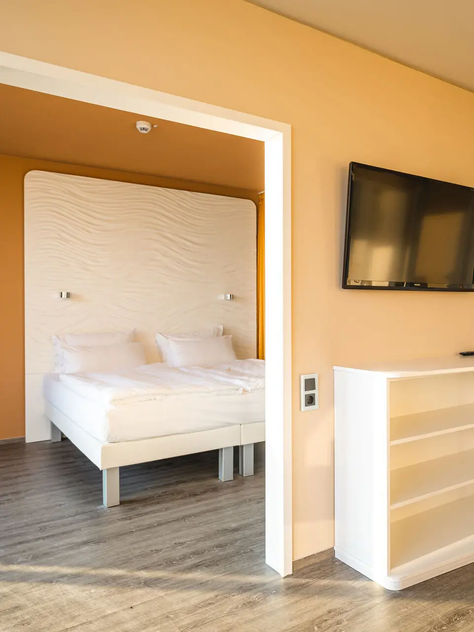 Ein Ausschnitt von einem Hotelzimmer mit Holzboden in dem ein weißes Regal über dem ein Fernseher hängt steht und einer Durchgangstür, die in das Schlafzimmer mit senfgelben Wänden und einem weißen Bett führt.