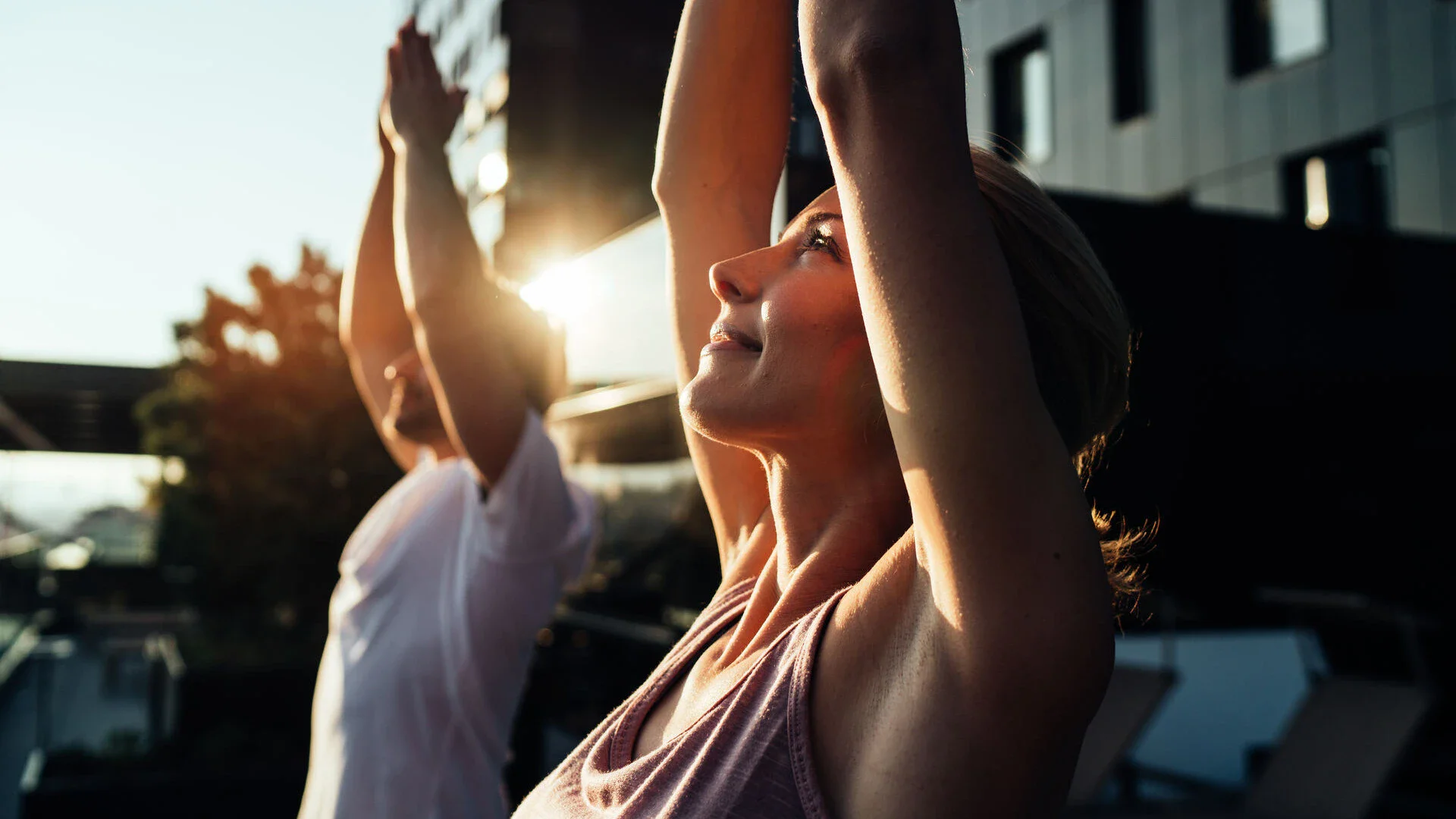 Zwei Personen stehen auf einer Außenfläche und machen Yoga. Durch sie hindurch scheint die strahlende Morgensonne.