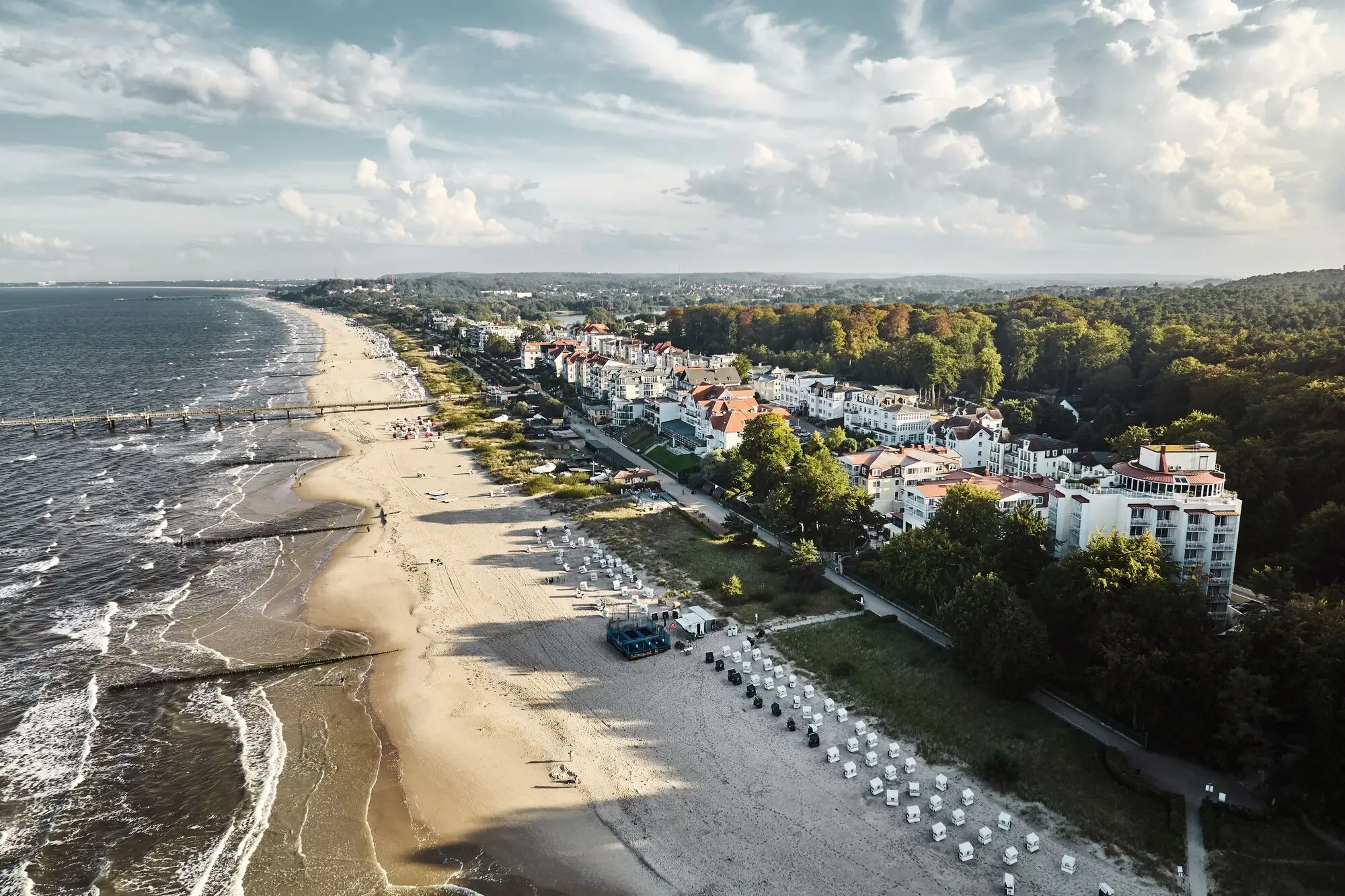 aja Bansin Ein Strand mit Gebäuden und Bäumen, unter einem bewölkten Himmel, mit Blick auf das Meer und umliegende Landschaft aus der Vogelperspektive.