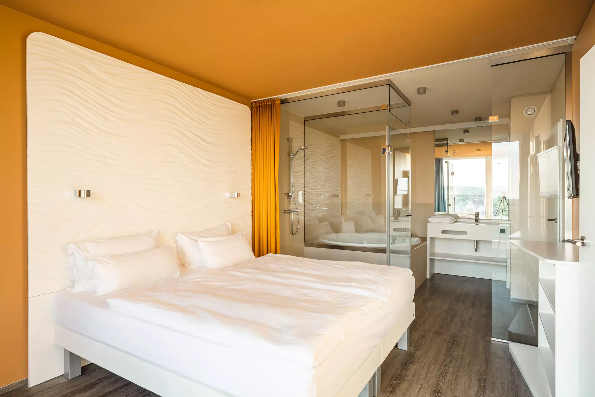 Hotelzimmer im aja Warnemünde Ein mit weißer Bettwäsche bezogenes Doppelbettsteht vor einer weißen Rückwand mit Struktur. Umrahmt von einer orangenen Wand und einem orangenen Vorhang steht das Bett in einem Raum mit orangefarbener Decke und dunklem Holzboden. Im Hintergrund ist hinter einer gläsernen Trennwand ein Badezimmer zu erkennen.