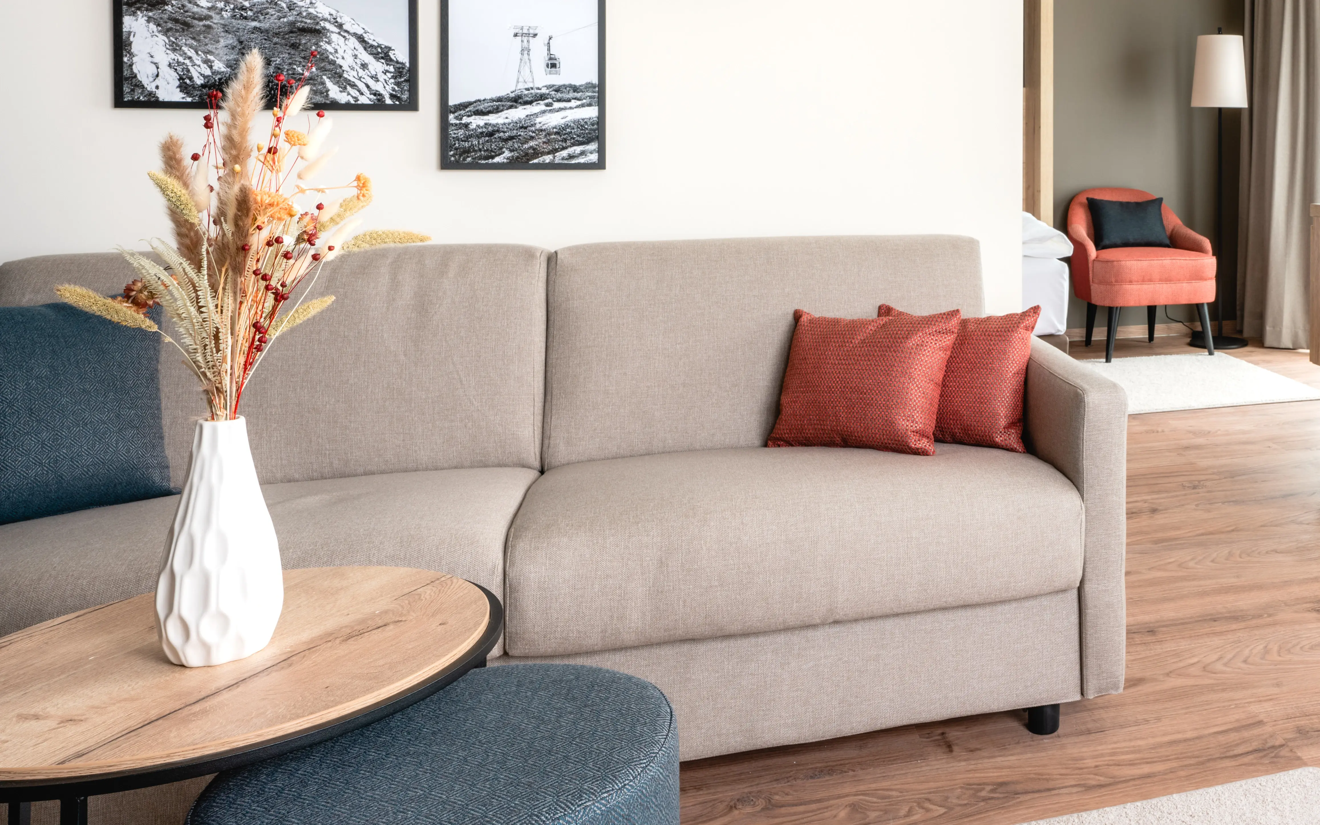 Modern eingerichtetes Hotelzimmer mit einem gemütlichen Wohnbereich. Ein beiges Sofa mit roten und blauen Kissen steht neben einem kleinen Tisch mit einer Vase voller Trockenblumen. An der Wand hängen schwarz-weiße Landschaftsfotografien. Der Essbereich mit roten Stühlen befindet sich neben einer großen Fensterfront, die viel Tageslicht hereinlässt.