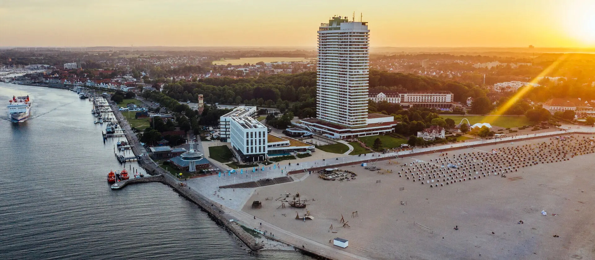 Eine Luftansicht einer Promenade mit Gebäuden, einem hohen weißen Hotel und breitem Sandstrand. Aus der oberen Ecke scheint die untergehende Sonne warm in das Bild.