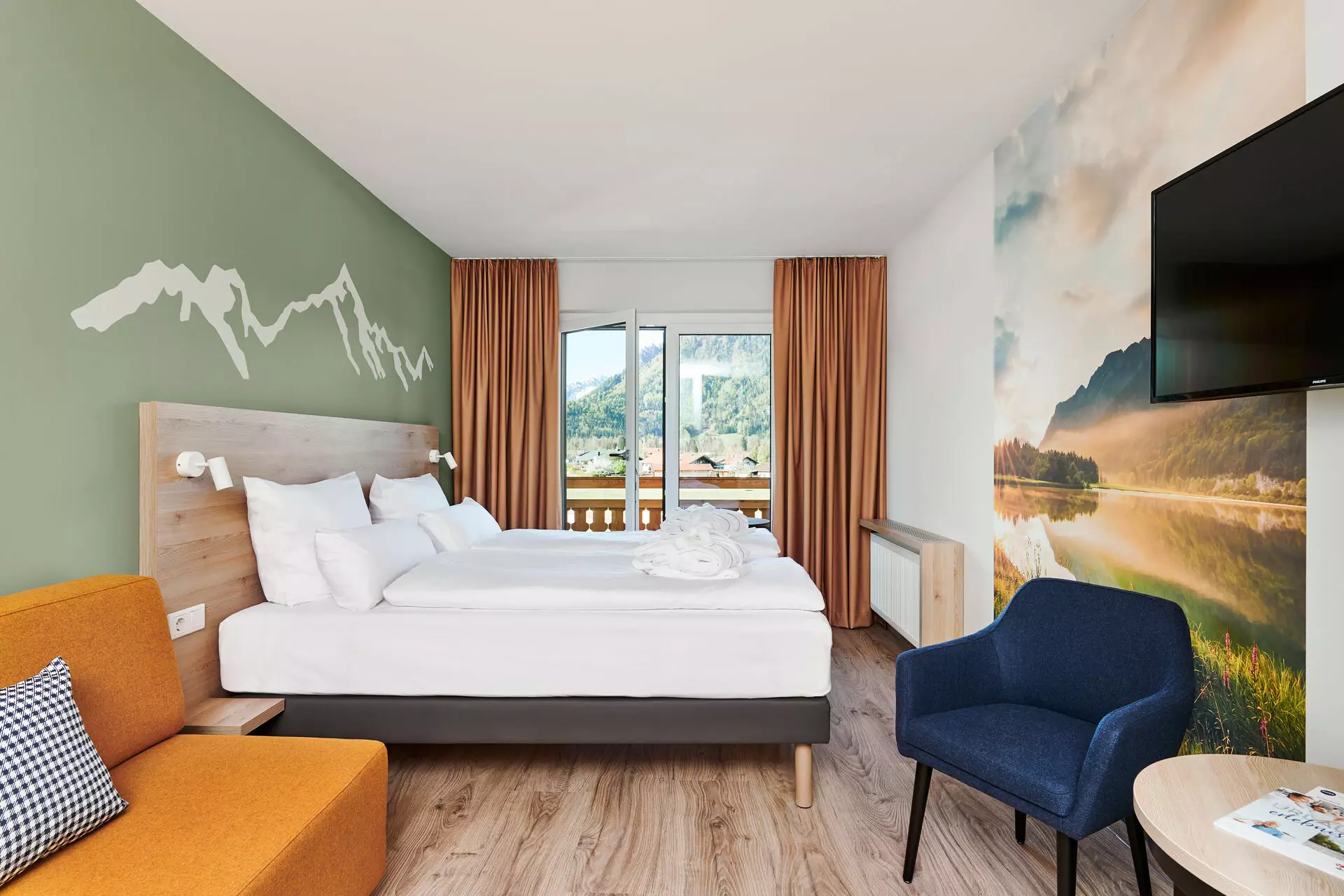 Hotelzimmer im aja Ruhpolding In einem hellen Raum steht ein mit weißer Bettwäsche bezogenes Doppelbett mit einem hölzernem Kopfteil vor einer sanft-grünen Wand. Auf der grünen Wand ist in weiß die Skyline von Berggipfel gemalt. Im Hintergrund ist ein bodentiefes Fester eingerahmt von hellbraunen Vorhängen geöffnet mit Blick auf die Berge. Die rechte Wand des Raums ist mit einer Fototapete mit Bergsee-Motiv verziert. Davor steht ein dunkelblauer Stuhl mit hohen Armlehnen. Vorne links sind noch die Ansätze eines orangefarbenen Sofas zu erkennen, auf dem ein schwarz-weiß kariertes Dekokissen platziert ist.