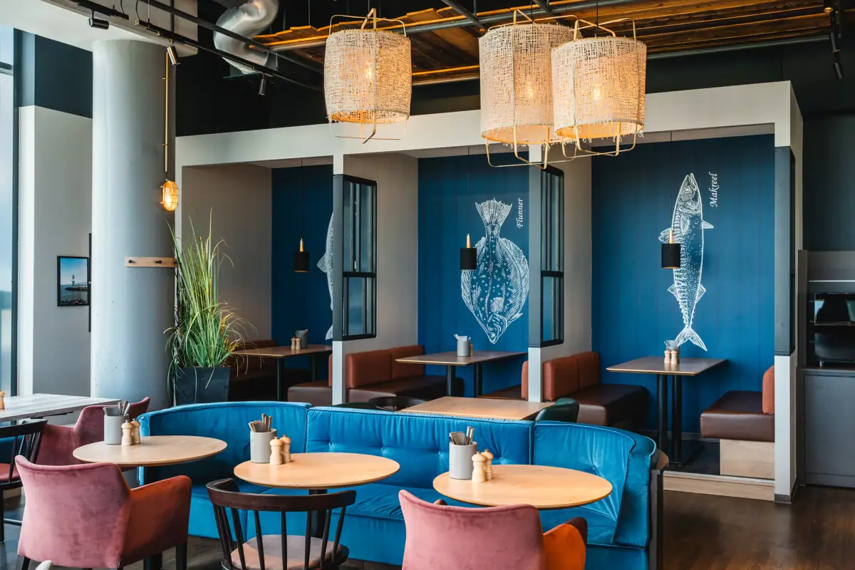 Restaurant im aja Warnemünde Ein Raum mit Sitznischen, maritim gestaltet und mit Tischen und Stühlen eingerichtet.