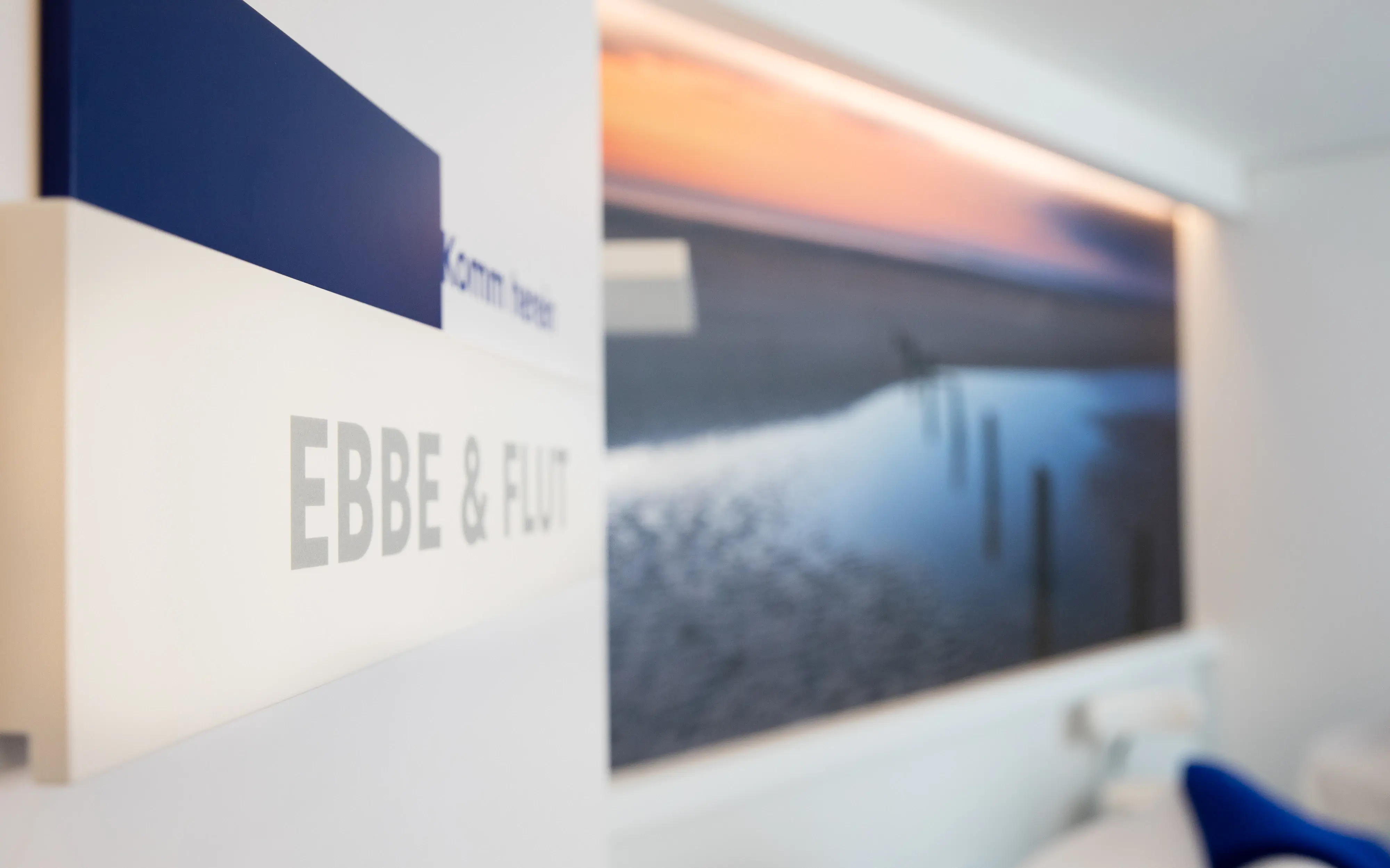Detailaufnahme eines Schildes mit der Aufschrift 'Ebbe & Flut' vor einem maritimen Wandbild mit Sonnenuntergang und Meeresszene.