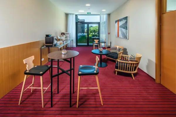 Medialounge Ein Raum mit rotem Teppich, verteilten Sitzmöglichkeiten, einer Kaffeebar und einer großen Terrassentür im Hintergrund.