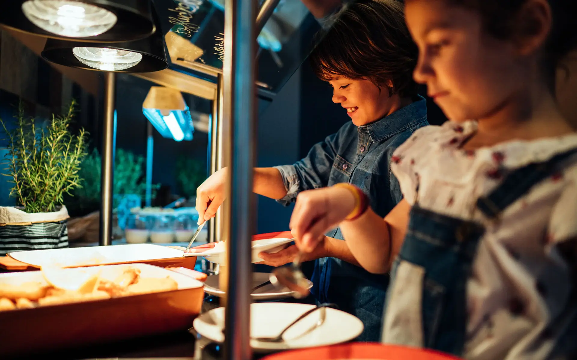 Kinderbuffet Zwei Kinder stehen an einem gedämpft beleuchteten Kinderbuffet und bedienen sich an warmen Speisen.