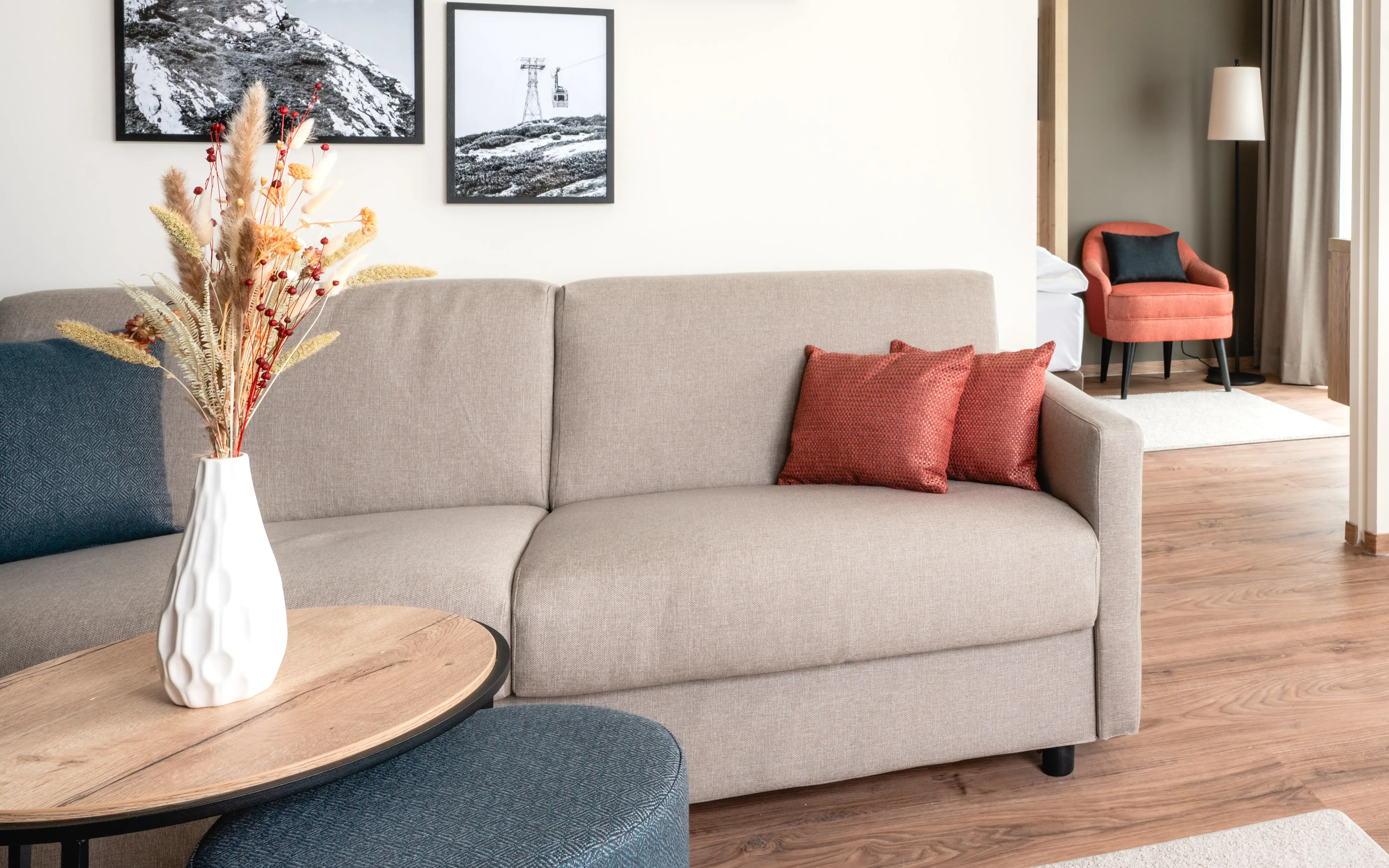 Ein helles und modernes Hotelzimmer mit separatem Wohn- und Essbereich. Ein bequemes Sofa mit farblich abgestimmten Kissen steht vor einer Wand mit schwarz-weißen Landschaftsbildern. Daneben steht ein Tisch mit roten Stühlen.Große Fenster mit Vorhängen lassen viel Tageslicht herein. Im Hintergrund ist der Schlafbereich mit einem weiteren Sessel sichtbar.